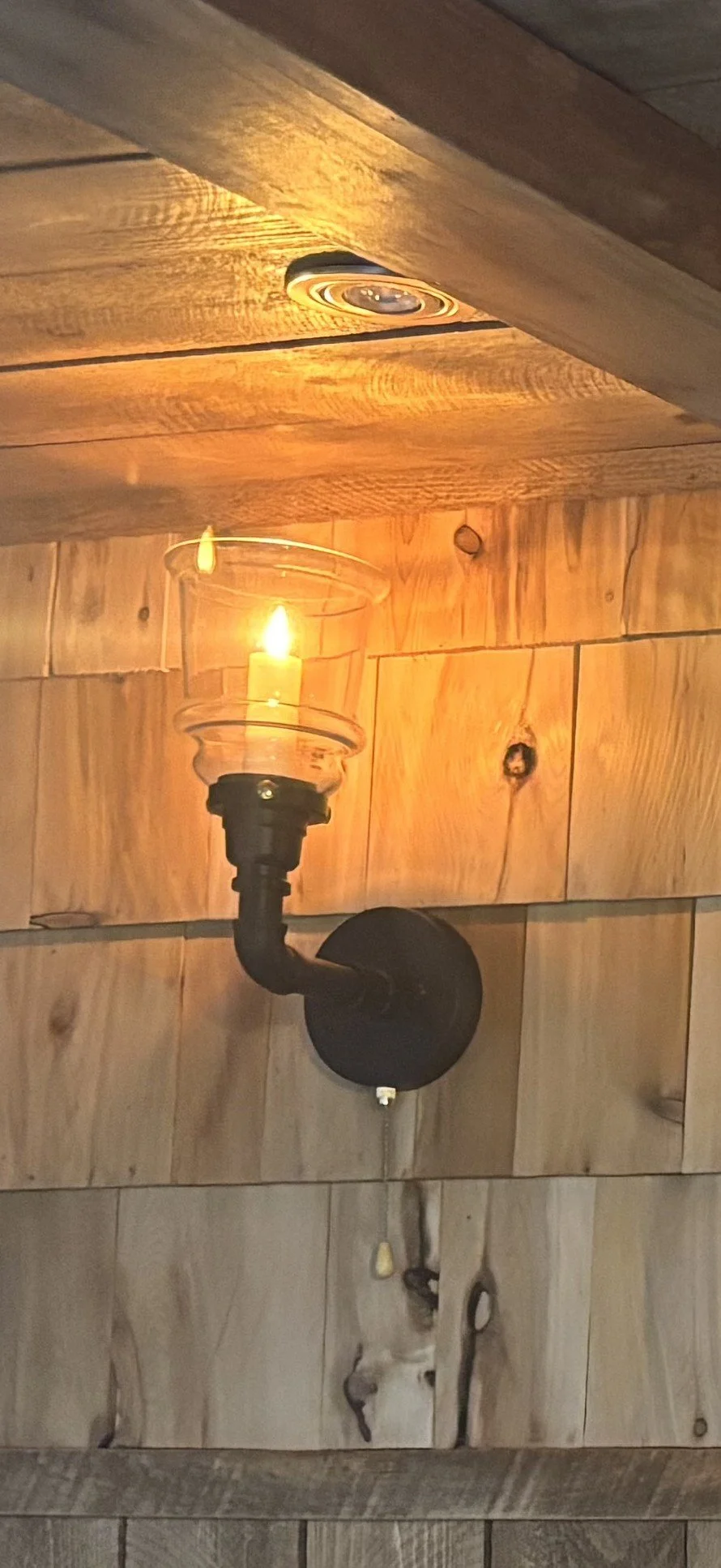 Wall-mounted black lamp with exposed bulb and glass cover, casting a warm glow on wooden wall panels.