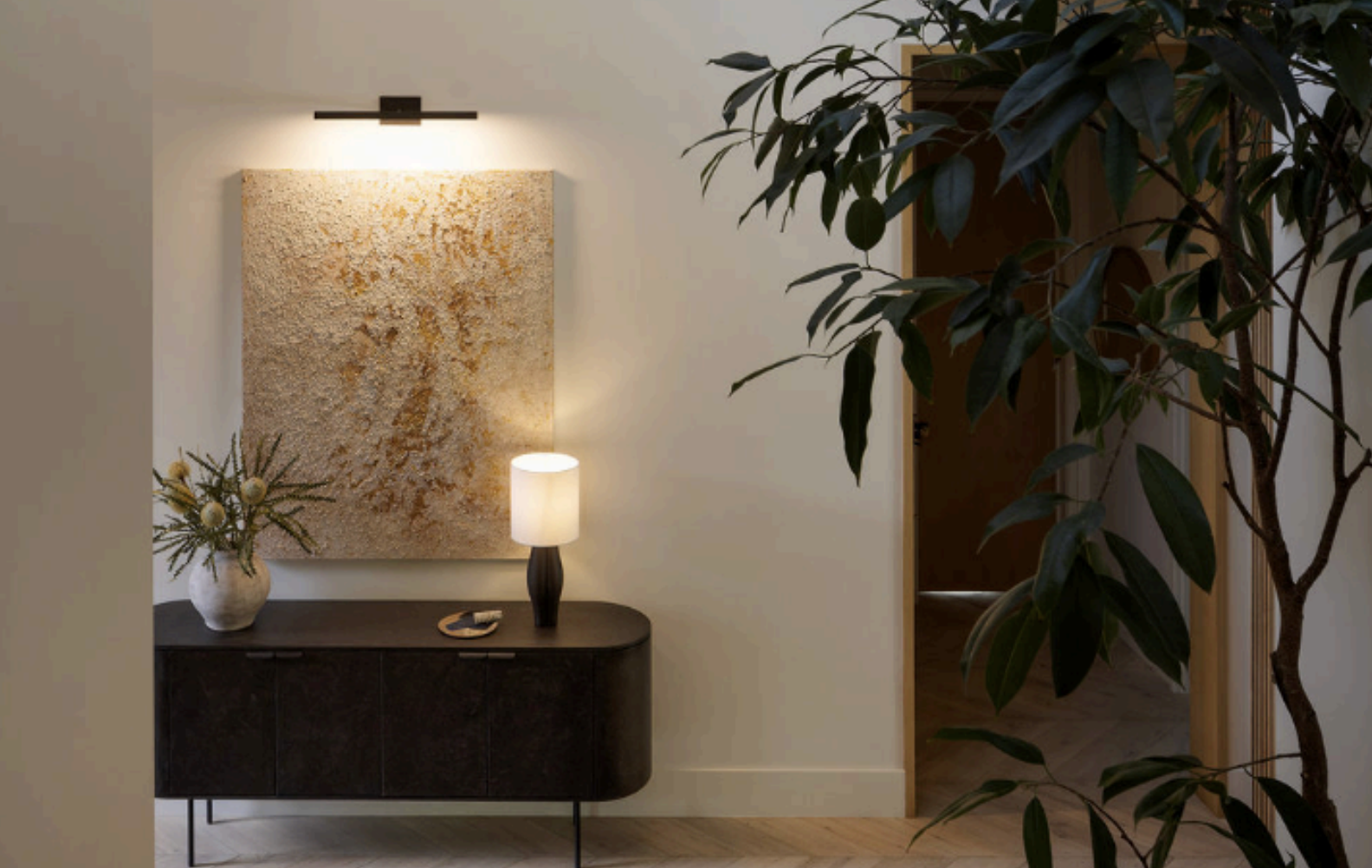 Interior of a modern home hallway with a black console table, a decorative vase with greenery, a table lamp, a textured wall art, and a large indoor plant in the foreground. Luxury art staging in Los Angeles featuring museum-quality contemporary 