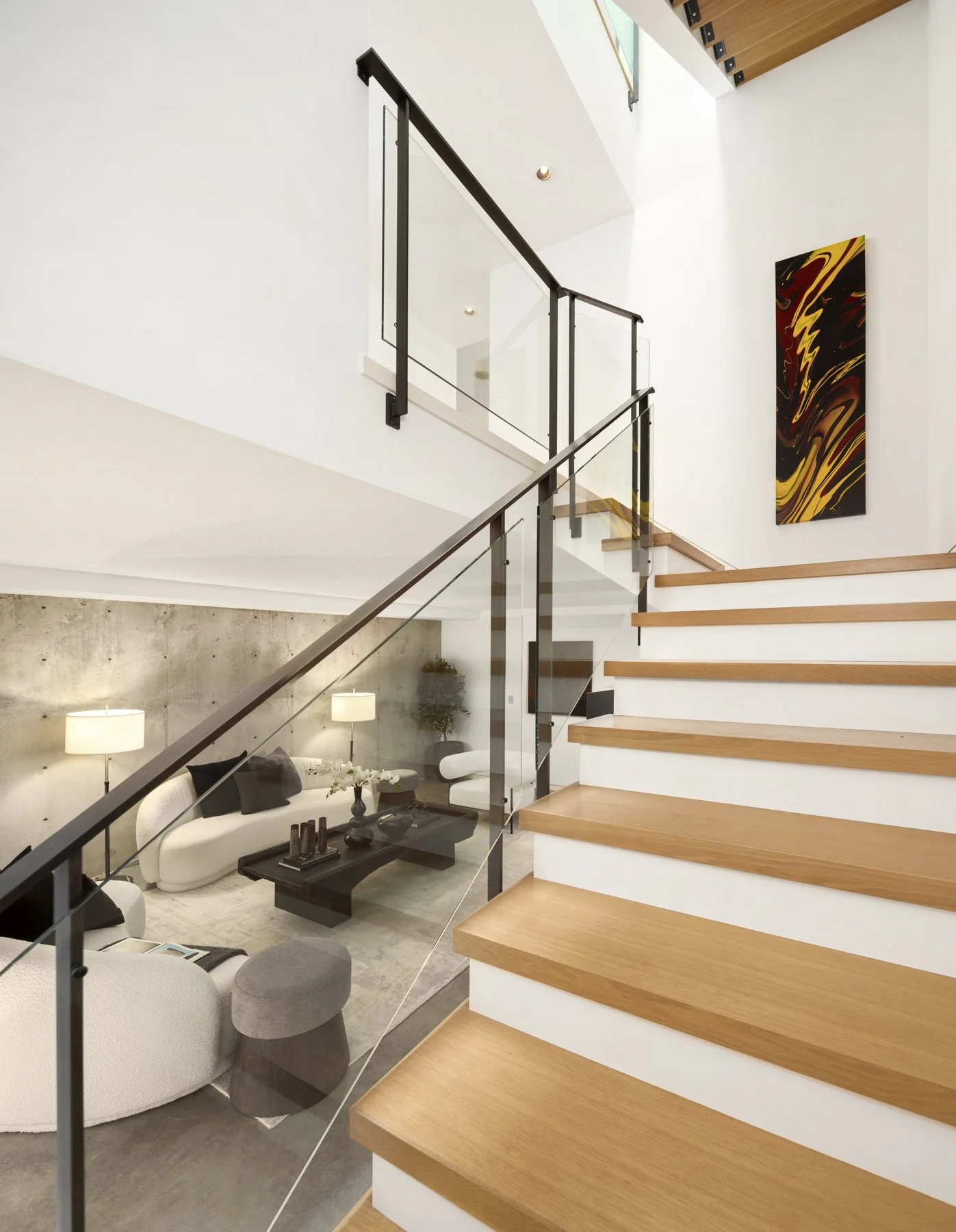 Interior view of a modern home with wooden staircase, white walls, and a glass railing. A living room area with a white sofa, black coffee table, and wall art visible in the background.