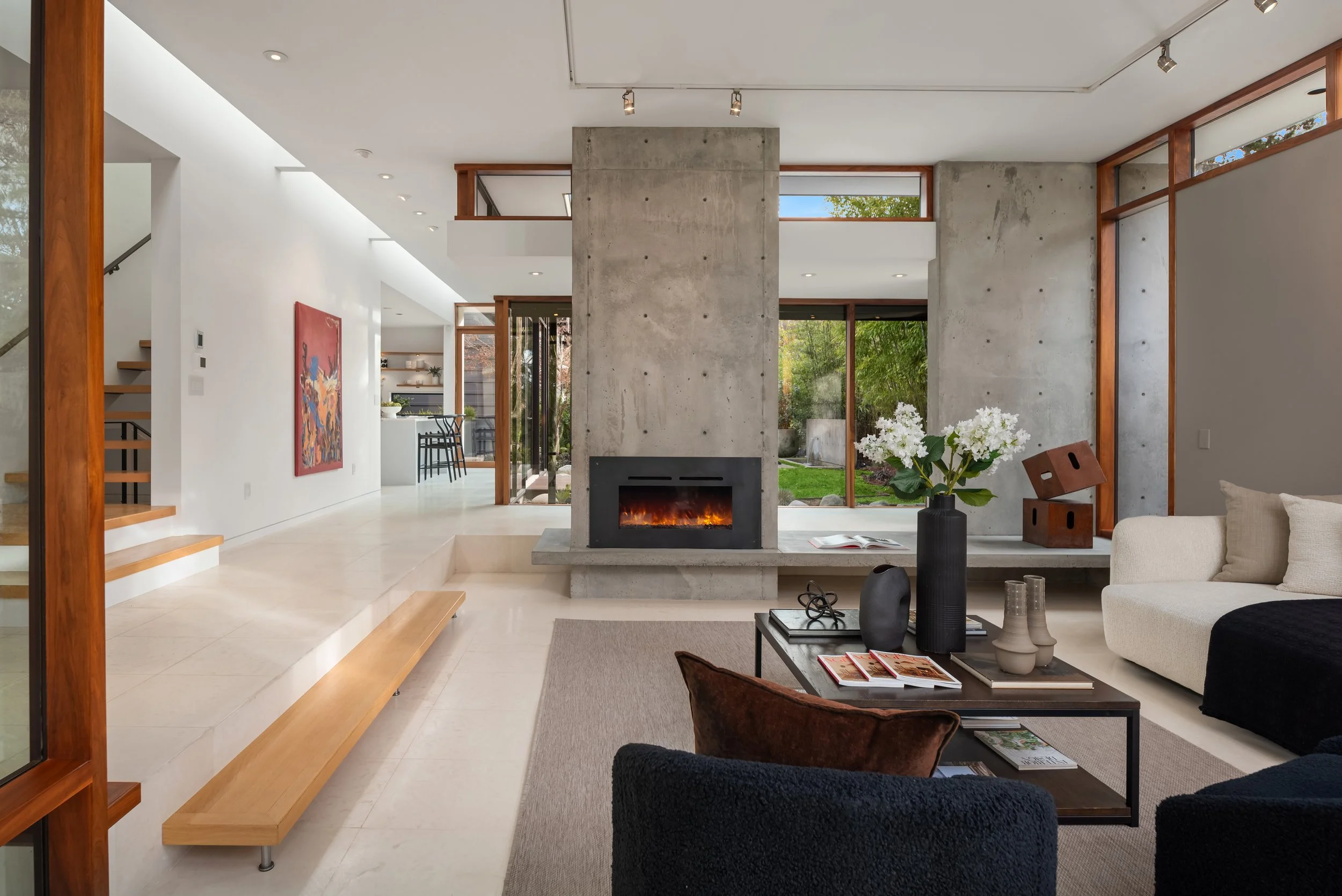 Modern living room with concrete fireplace, large windows, white walls, wooden accents, and furniture including a black armchair, vases, and a sofa with pillows.