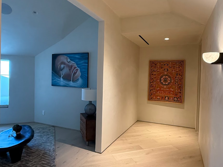 Interior view of a home with a hallway and a living room. The hallway features a decorative wall hanging, while the living room has a large painting of a person's face sleeping, a side table with a lamp, a window, and a rug. Luxury art staging 