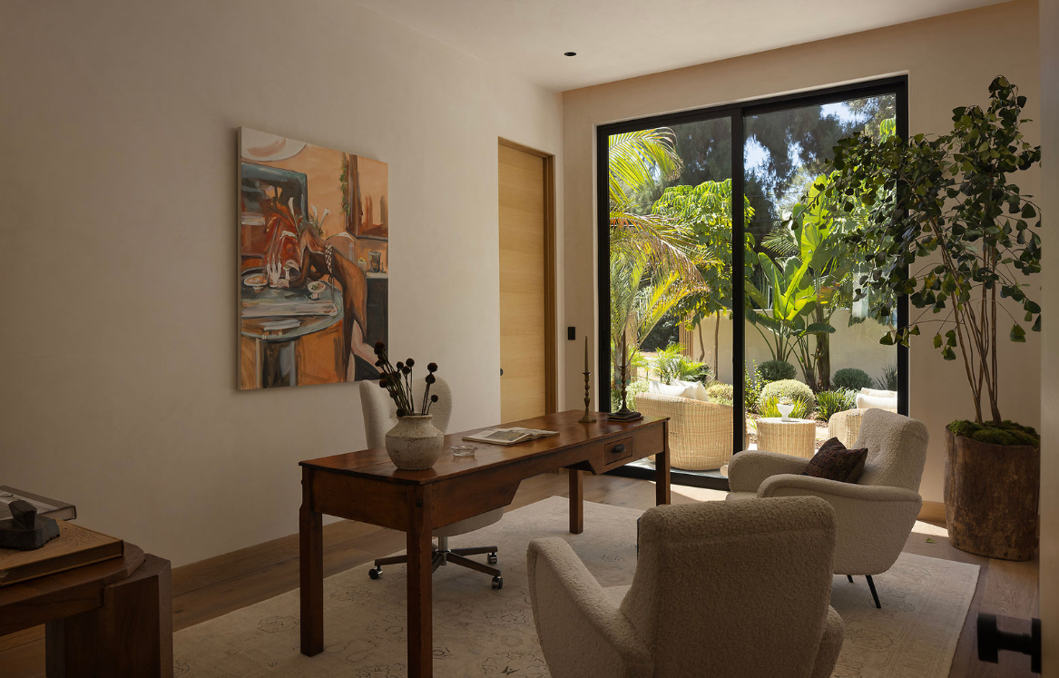 Living room with a wooden desk, two beige armchairs, and a large sliding glass door leading outside to a lush garden with trees and outdoor seating.