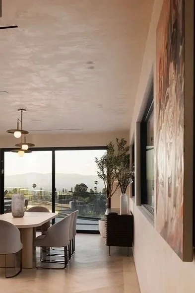 Interior of a modern dining room with a round table, six chairs, a large window showing a view of trees and water, a ceiling fan with lights, potted plant, and artwork on the wall.