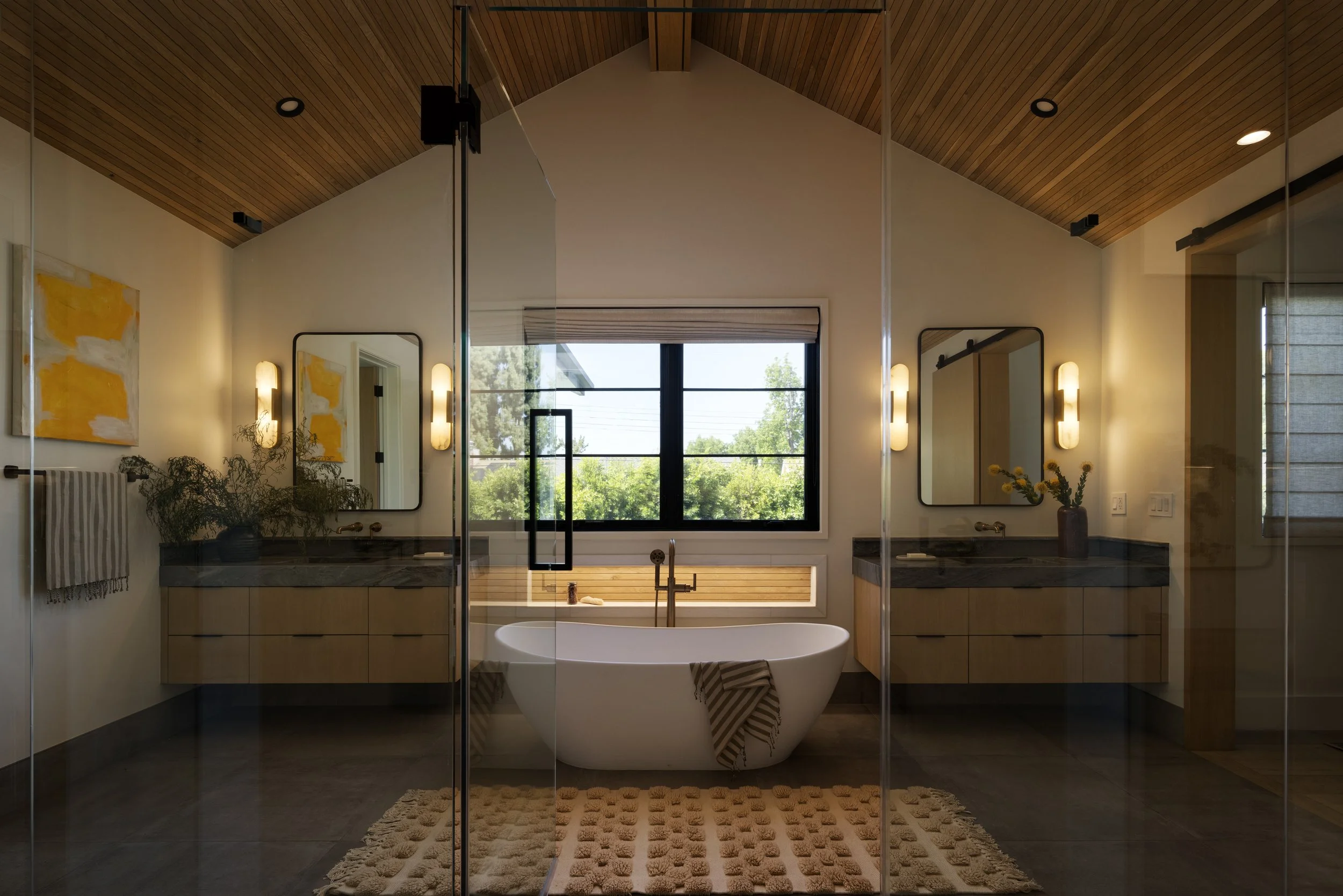 Modern bathroom with a large soaking bathtub in front of a window with trees outside. The bathroom features wooden ceiling, wall-mounted lights, mirrors, and a beige rug on dark tile floor.