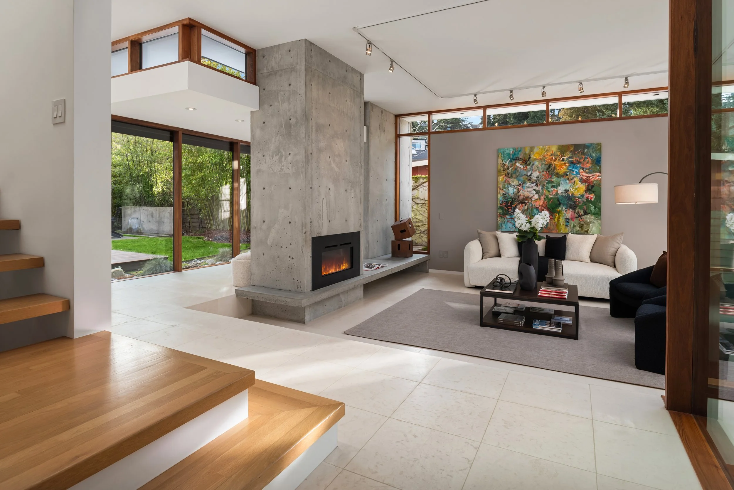 Modern living room with large windows, concrete fireplace, abstract painting, white sofa, black armchair, coffee table with decorative items, and hardwood stairs.