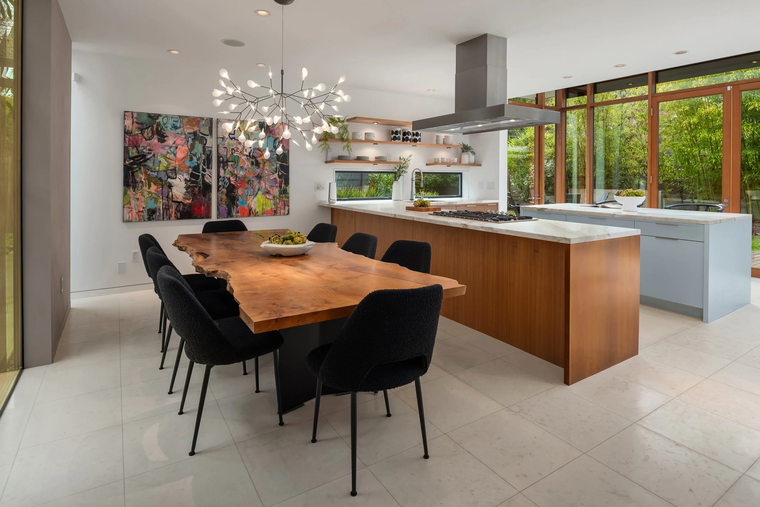 Modern kitchen with a wooden dining table and black chairs, open shelves with dishes, large windows with greenery outside, ceiling light fixture, and marble kitchen island.