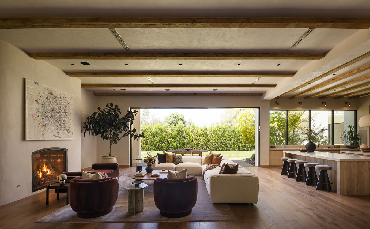 Interior of a modern house with wood-paneled ceiling and walls, a dining table with wine glasses, a colorful painting on the wall, and lush greenery visible through the windows.