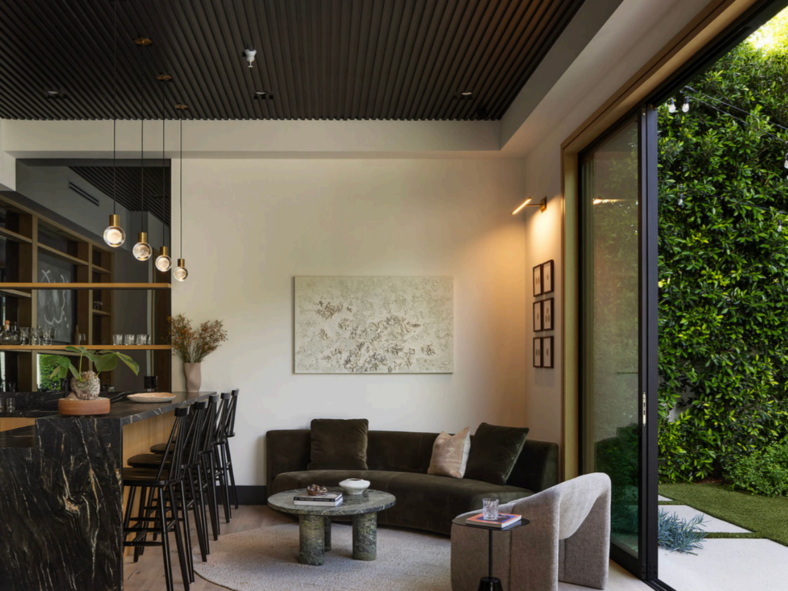 Modern living room with dark sofa, beige armchair, round marble coffee table, large window with greenery outside, wall art, and hanging pendant lights. Luxury art staging in Los Angeles featuring museum-quality contemporary artwork in a modern luxury