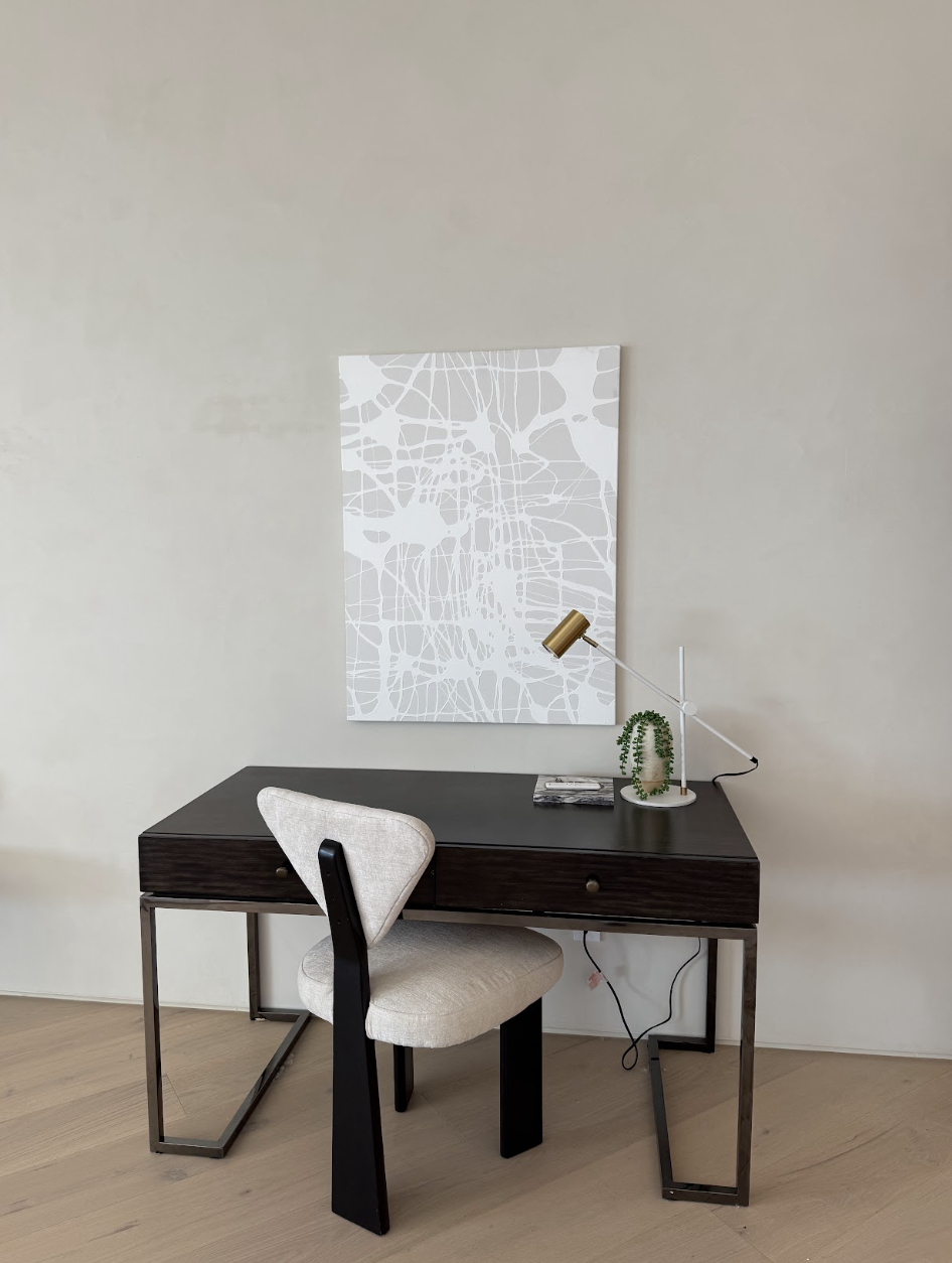 Modern home office with a dark wood desk, beige upholstered chair, white wall art, desk lamp, and a small green plant.
