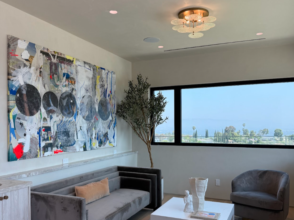 Living room with large abstract artwork on the wall, a gray sofa with a beige pillow, a small side cabinet, a white coffee table with decorative objects, a gray armchair, a tall potted plant, and a large window showing a landscape with trees and hills.