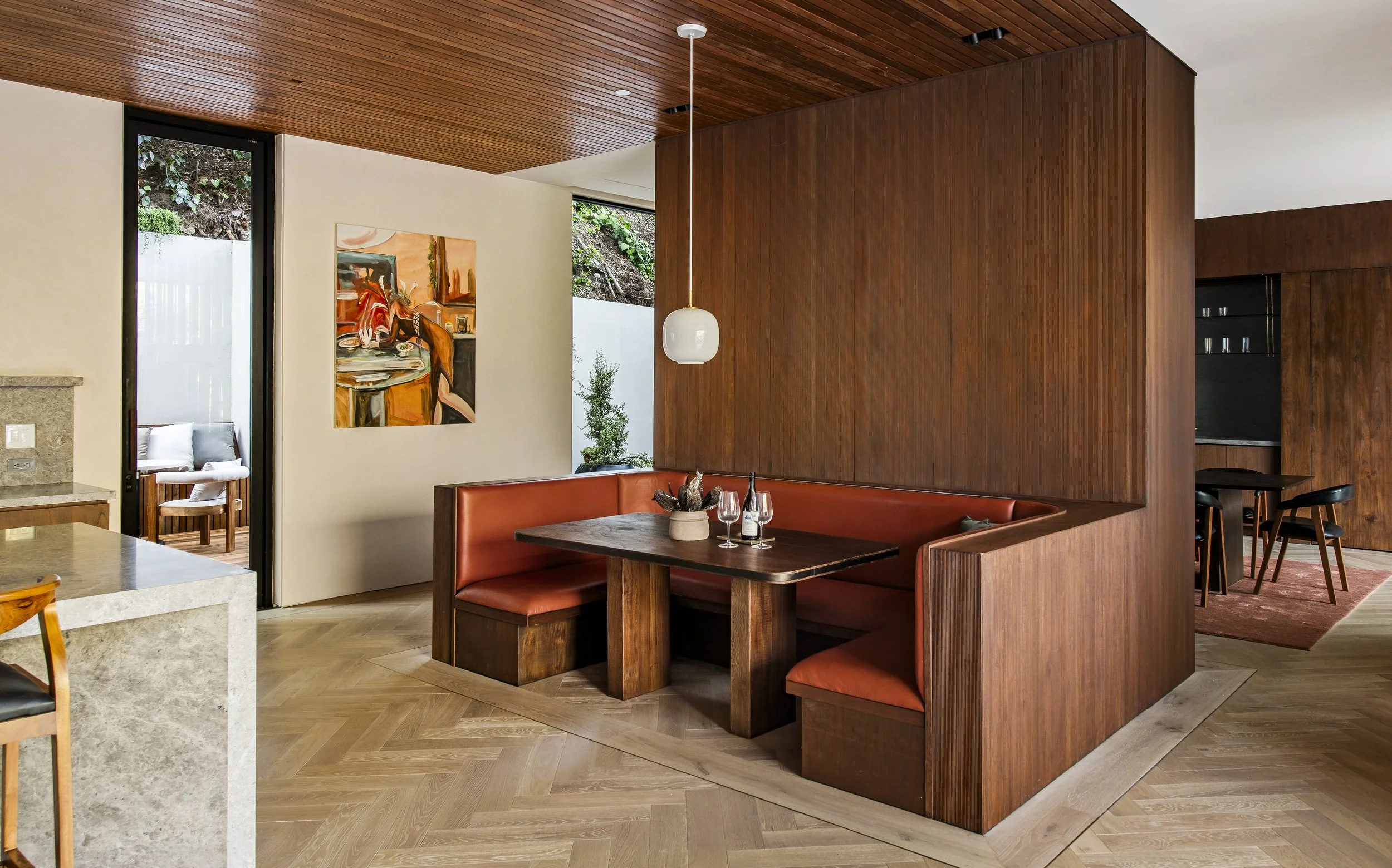 Interior of a modern dining area with wooden ceiling and wall, featuring a booth-style dining section with a wooden table, wine glasses, and a decorative centerpiece.