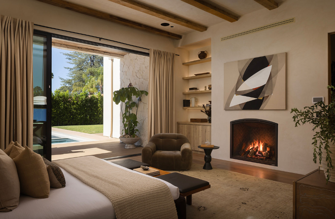 Living room with a fireplace, abstract wall art, large window with beige curtains, green plants, a cozy chair, and a view of an outdoor pool and garden.