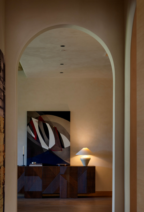 Interior view of a living room through an archway, featuring a wooden sideboard with geometric patterns, a table lamp, and an abstract painting on the wall.