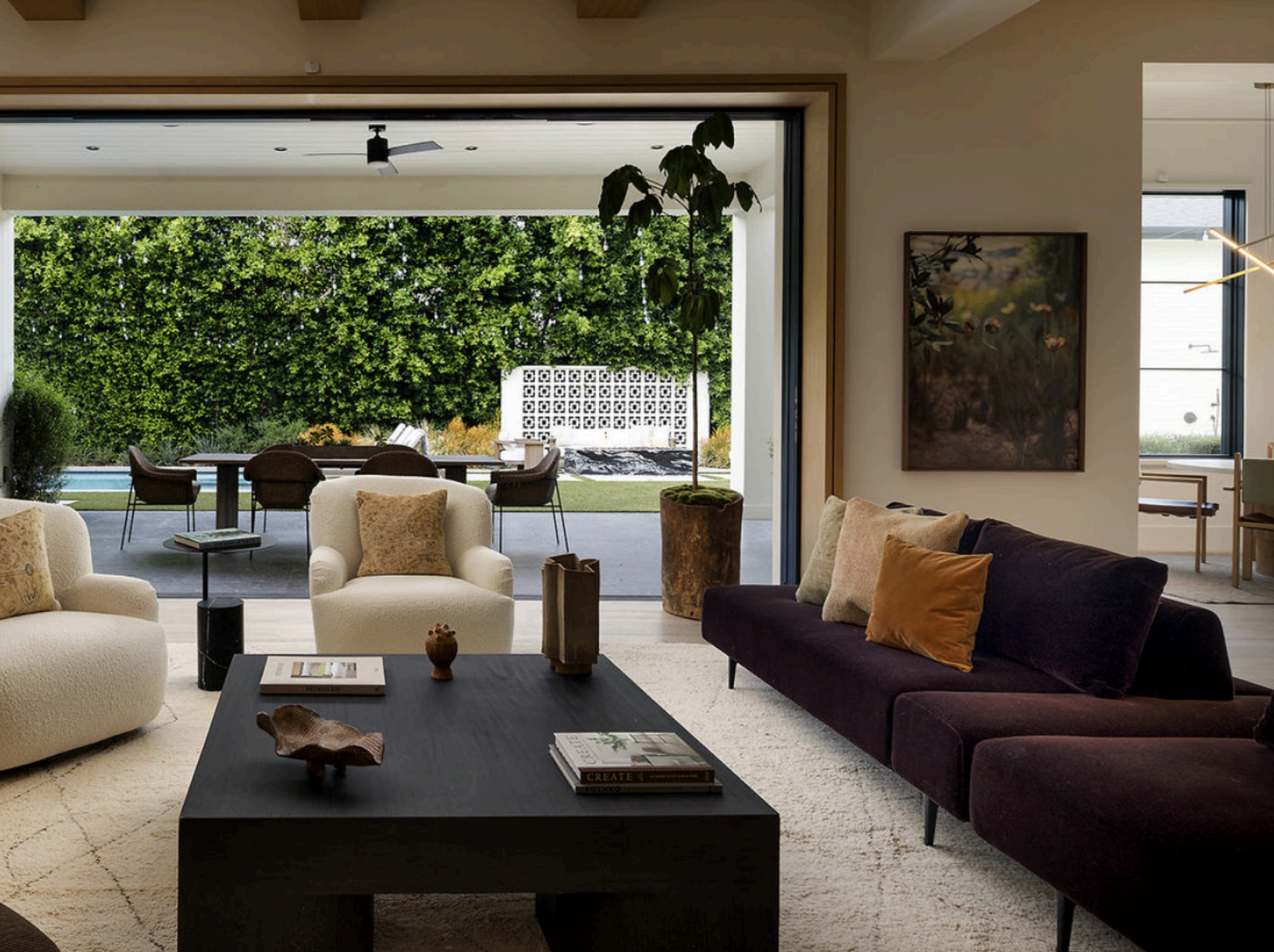 A modern living room with a wooden bookshelf filled with books and decorative items, a framed artwork of a woman's face submerged in water on the wall, a wooden dining table with a vase of flowers, a table lamp, and a chair, with a light-colored rug on a hardwood floor.