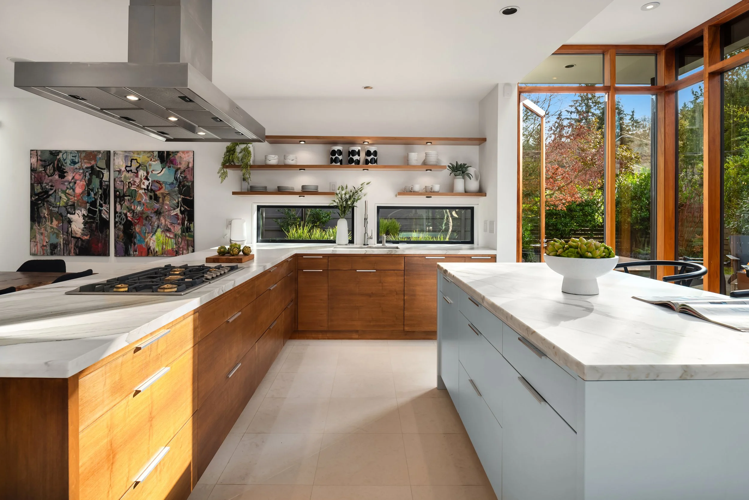 Modern kitchen with white and wood cabinets, open shelves with dishes, large windows with a view of trees, and a white marble island with a bowl of artichokes.