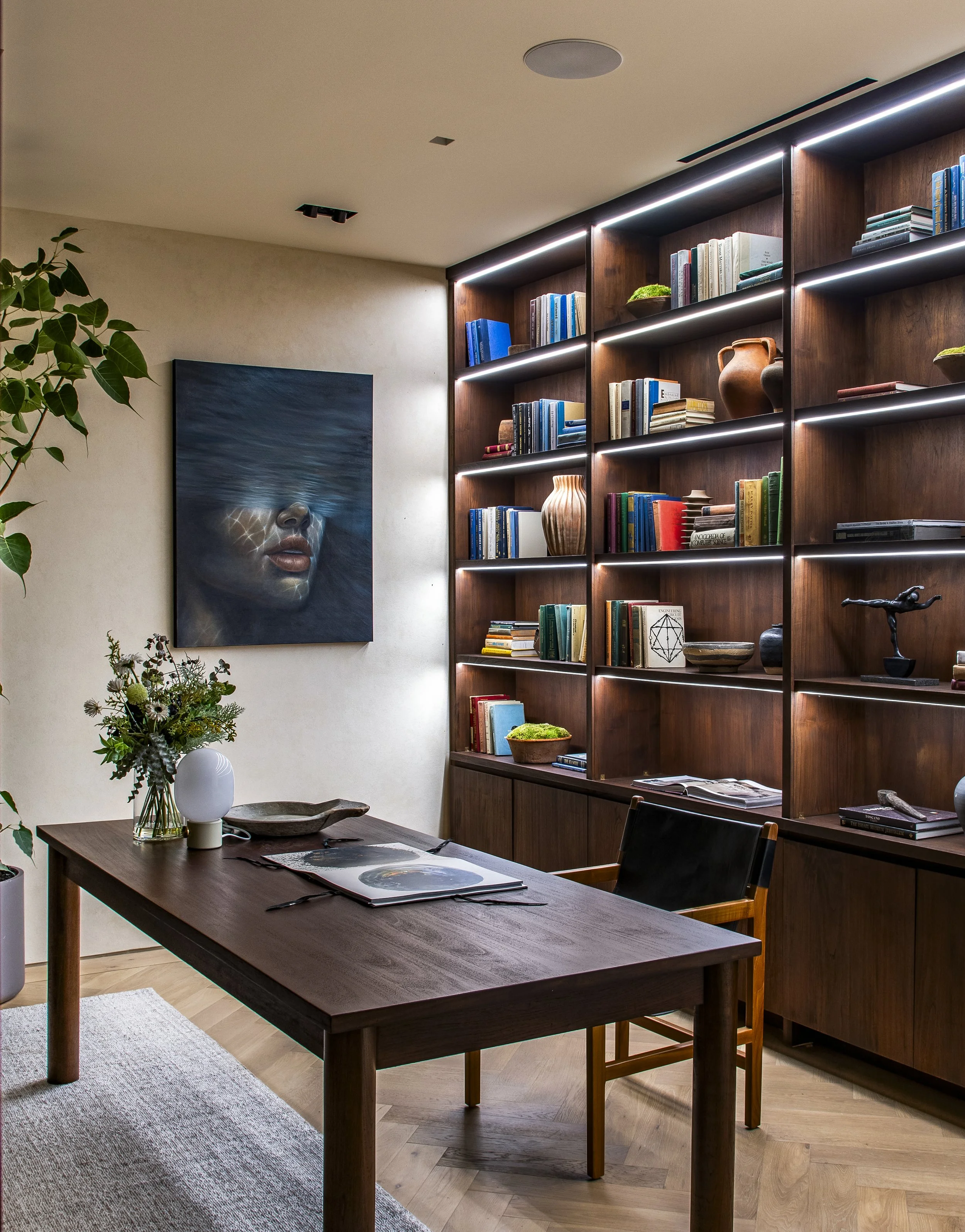 A modern dining room with a dark wooden table, a vase with flowers, and a textured beige rug. Behind, a large wooden bookshelf with LED lighting displays books and decorative items. On the wall, a artwork of a face submerged in water. There is a chair with wooden arms and black cushion at the table.