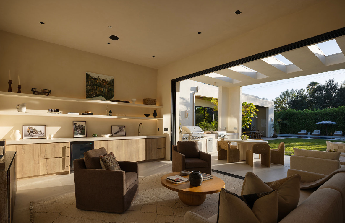Living room with brown and beige furniture, open to outdoor patio with grill and seating, natural light through skylights.