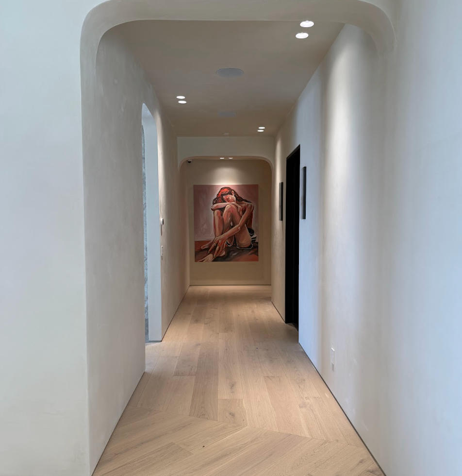 A hallway with cream-colored walls and light wood flooring, leading to a painting of a woman sitting with her knees drawn to her chest at the end of the hall. Luxury art staging in Los Angeles featuring museum-quality contemporary artwork in a modern