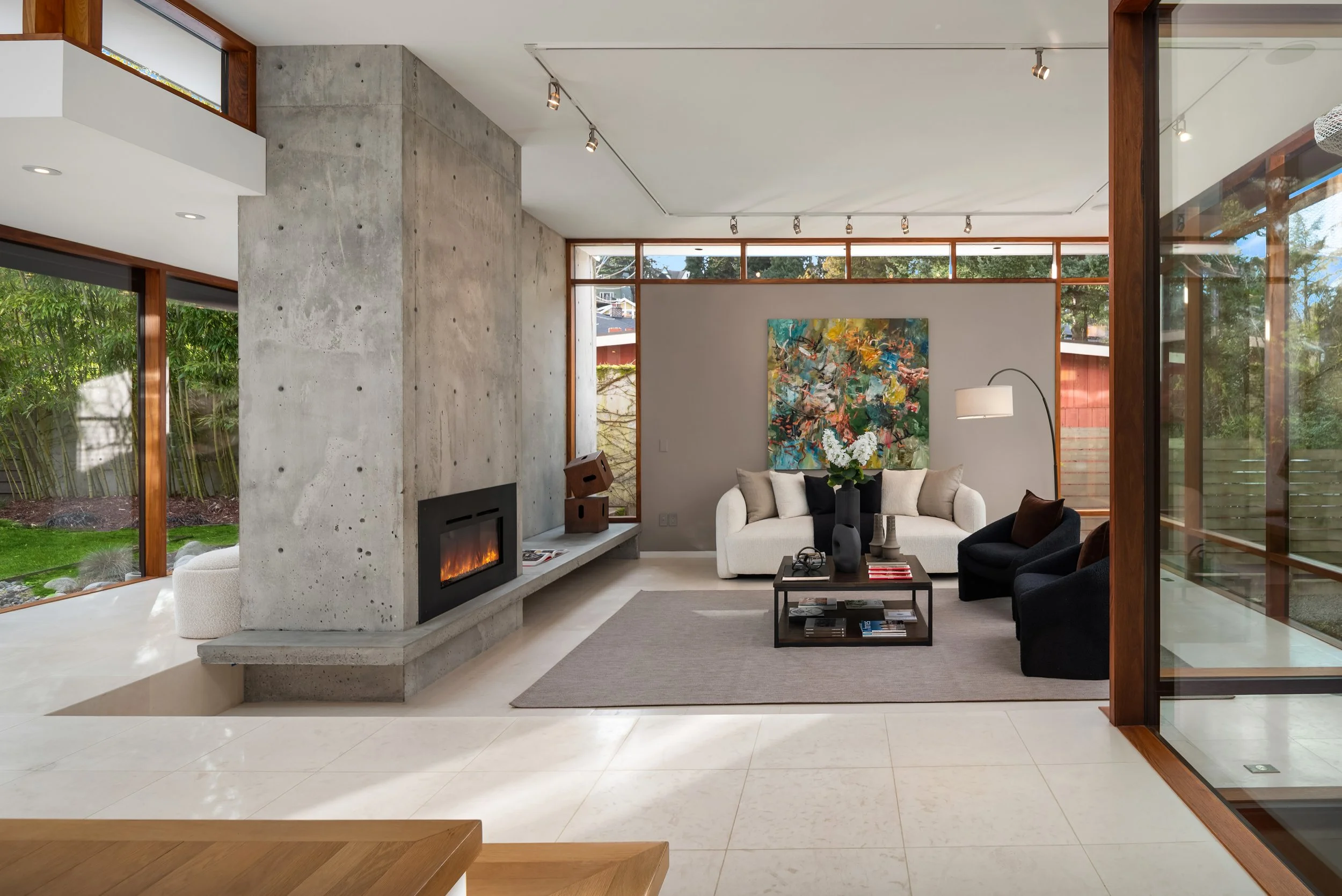A modern living room with large windows, a concrete fireplace, white sofa, black armchair, abstract painting, and a glass sliding door.