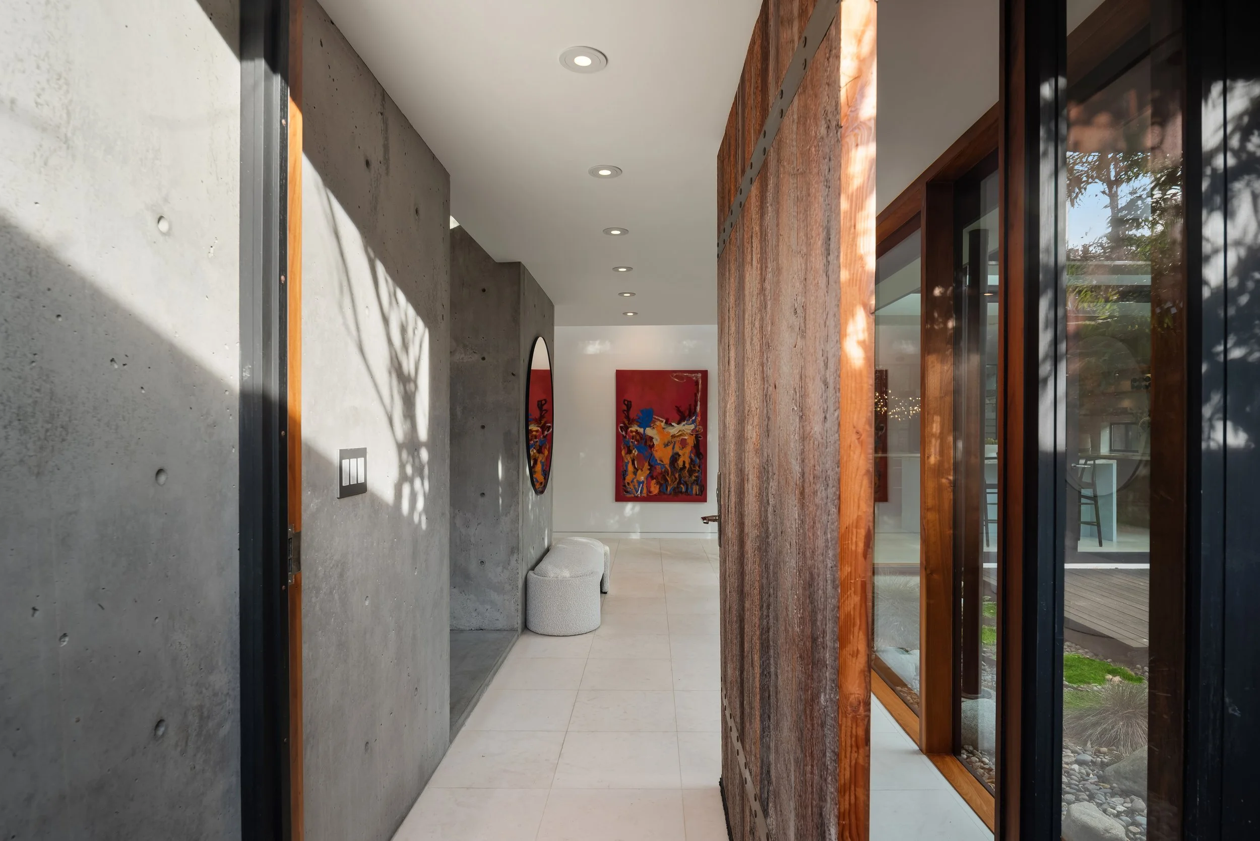 Interior of a modern home with wood-paneled ceiling, white walls, and a corner built-in dining booth with orange seating, a wooden table, and decorative items. Artwork on the wall and an open doorway leading to an outdoor area with greenery.