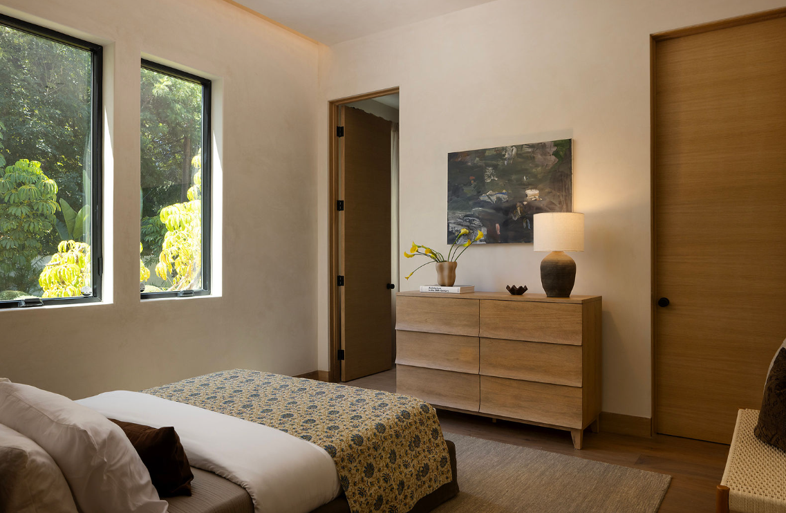 A cozy bedroom with two large windows showing green trees outside, a bed with white and brown pillows, a wooden dresser with a vase of flowers and a table lamp on top, and a piece of artwork hanging above the dresser.