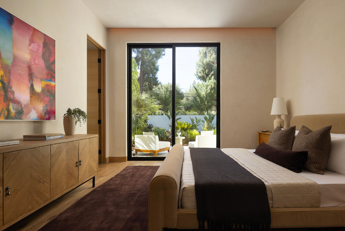Bedroom with a large bed, pillows, a nightstand with a lamp, a wooden dresser with books and a potted plant, and a balcony with outdoor seating and greenery.