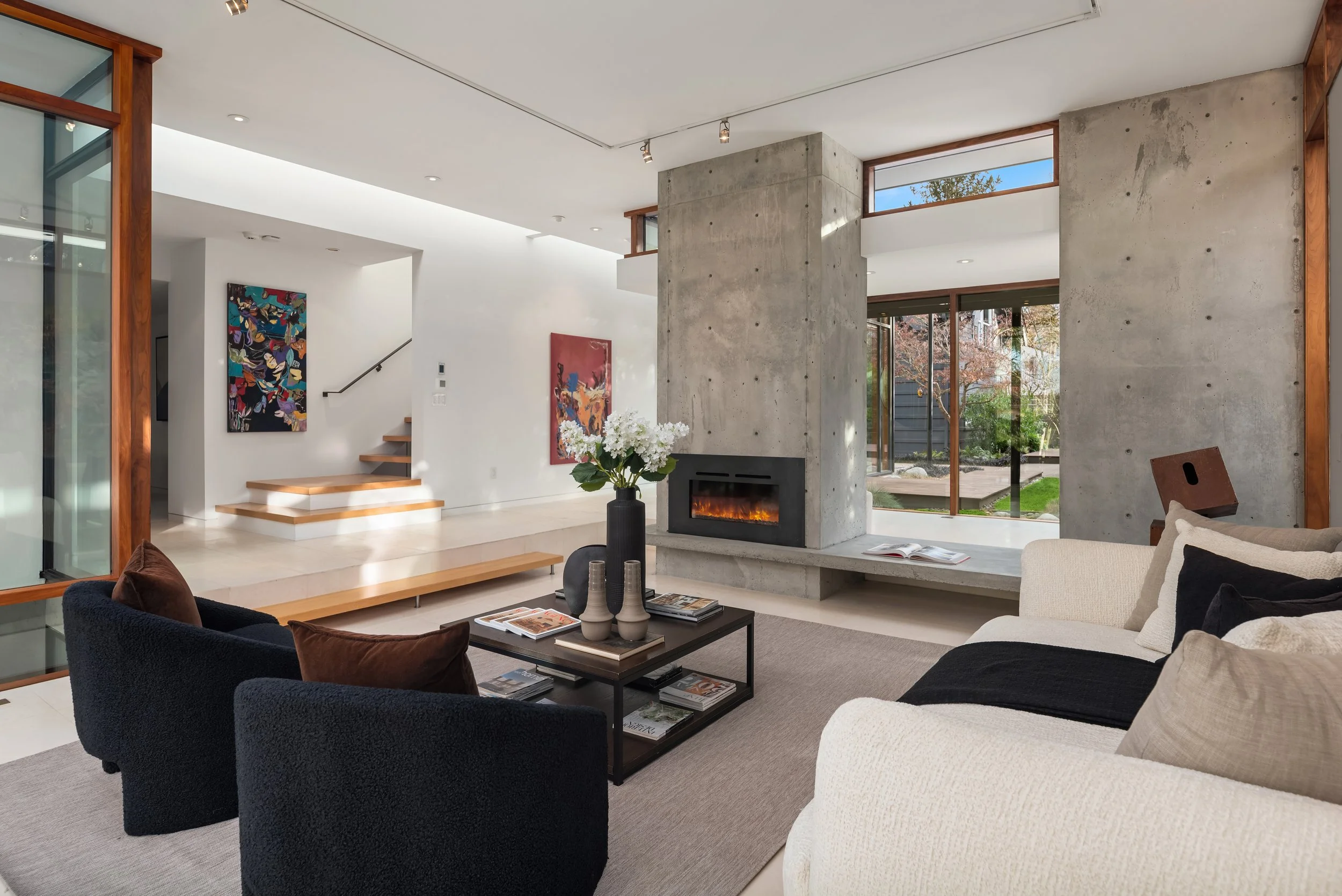 Modern living room with a concrete fireplace, large windows, art on white walls, and furniture including a white couch and black chairs.
