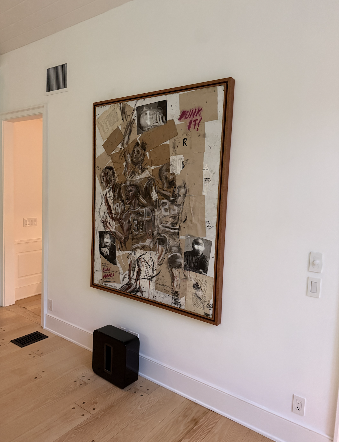 Interior of a modern home office or library with a wooden table, a bookshelf filled with books and decorative items, a large wall painting of a woman's face partly submerged in water, and a tall leafy plant.