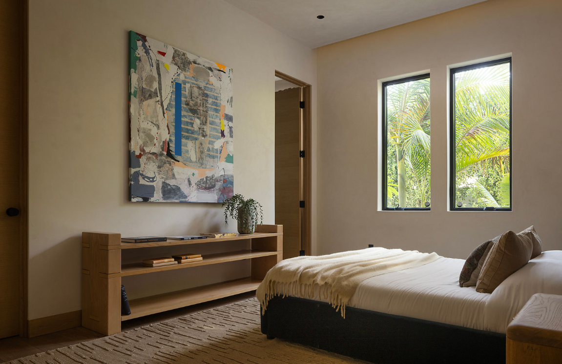 A bedroom with a bed, pillows, and a blanket, a wooden shelf with books and a plant, abstract artwork on the wall, and two windows showing greenery outside.