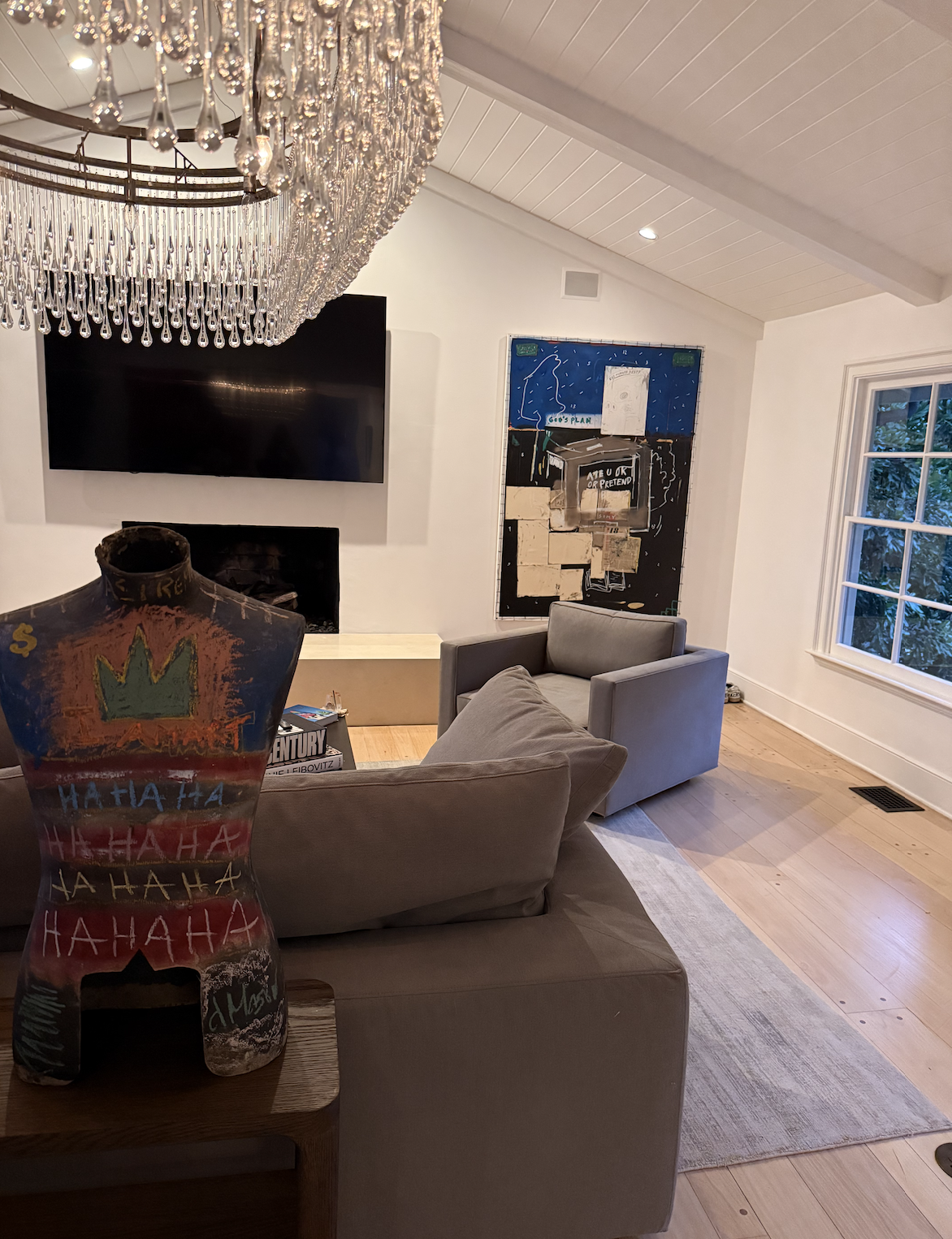 Interior of a modern house with wood-paneled ceiling and walls, a dining table with wine glasses, a colorful painting on the wall, and lush greenery visible through the windows.