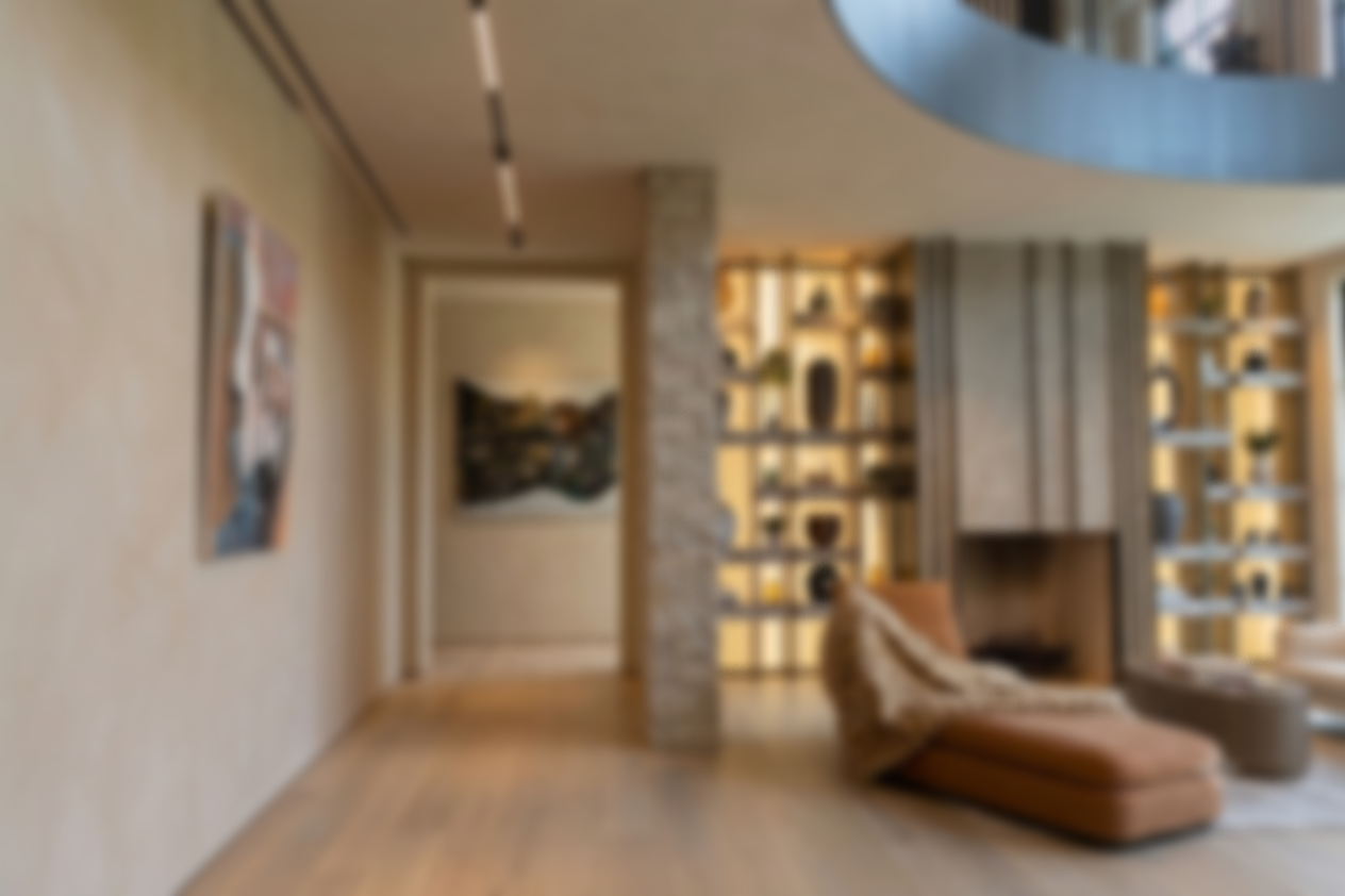 Blurred interior of a modern living room with artwork on the wall, a built-in bookshelf with decorative items, and a lounge chair near a large window.