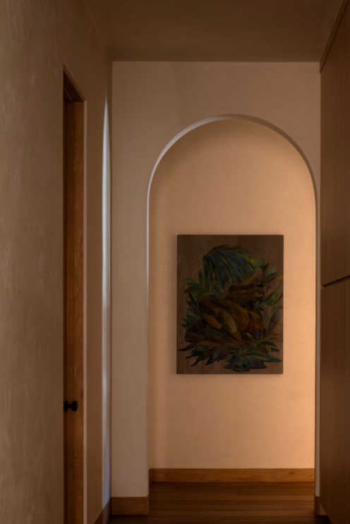 Interior view of a hallway with wooden flooring, a framed dark-colored botanical painting on the wall, and an arched opening leading to a lit room.