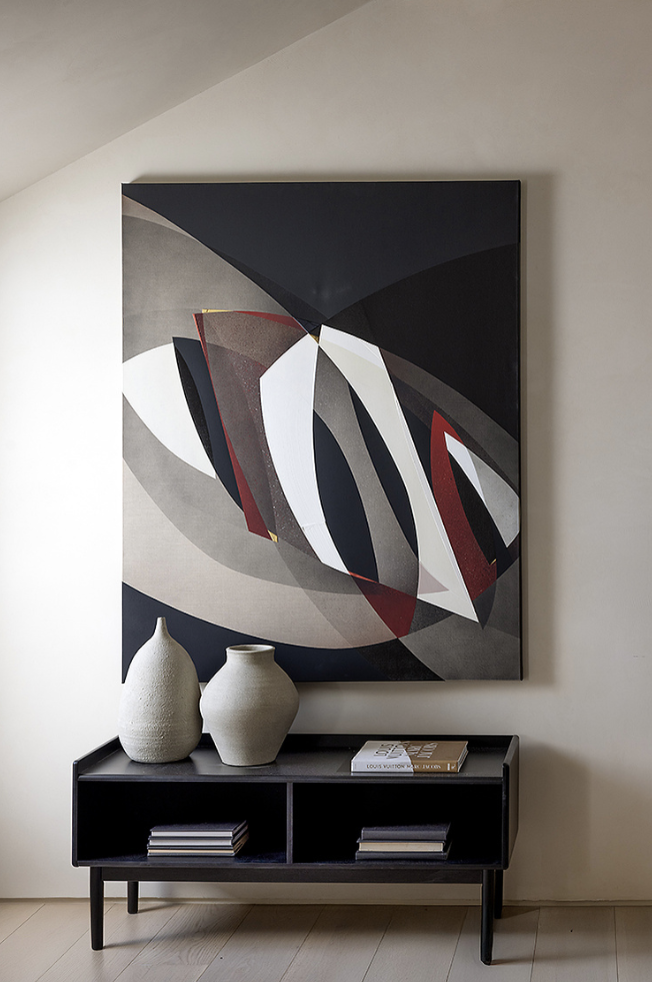 Abstract geometric painting with black, white, red, gray, and beige shapes hanging on a beige wall above a black console table with two large white vases and several books.