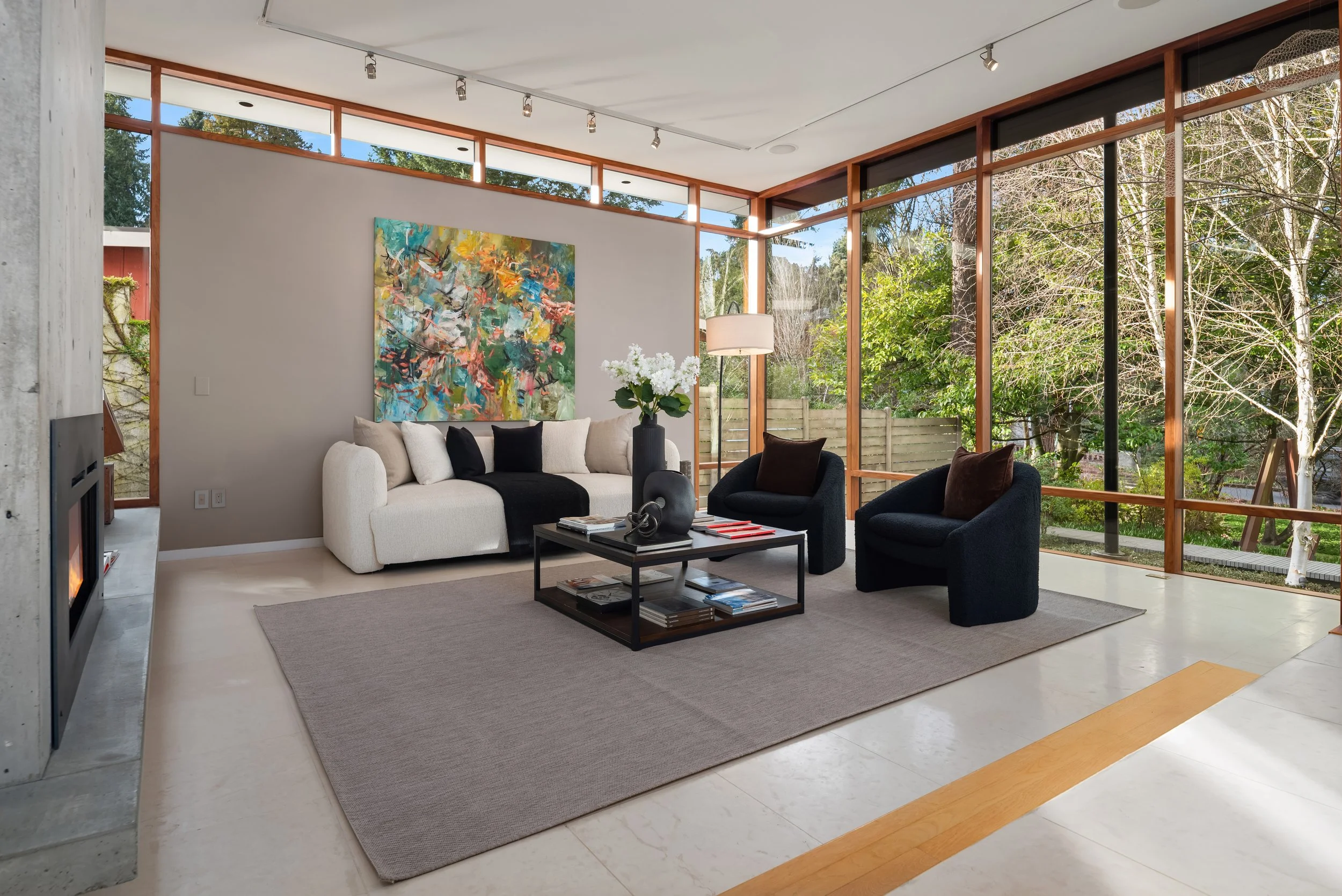 A modern living room or home office with a dark wooden dining table, a bookshelf with illuminated shelves filled with books and decorative items, a painting of a face partially underwater on the wall, and a vase with flowers on the table.