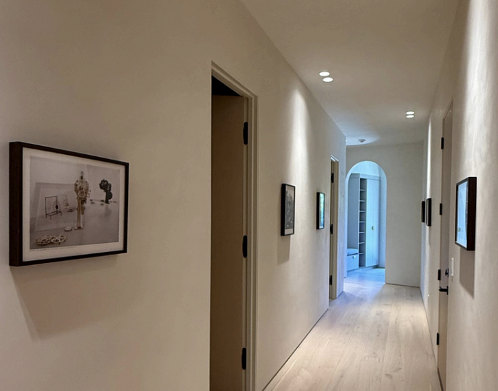A long, narrow hallway in an art gallery with framed artwork on both sides, angled wooden flooring, and a black ceiling with a skylight reflecting the space's interior.