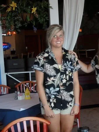 A young woman with short blonde hair wearing a black and white tropical patterned short-sleeve shirt and shorts, smiling in a restaurant with a table, chairs, and plants in the background.
