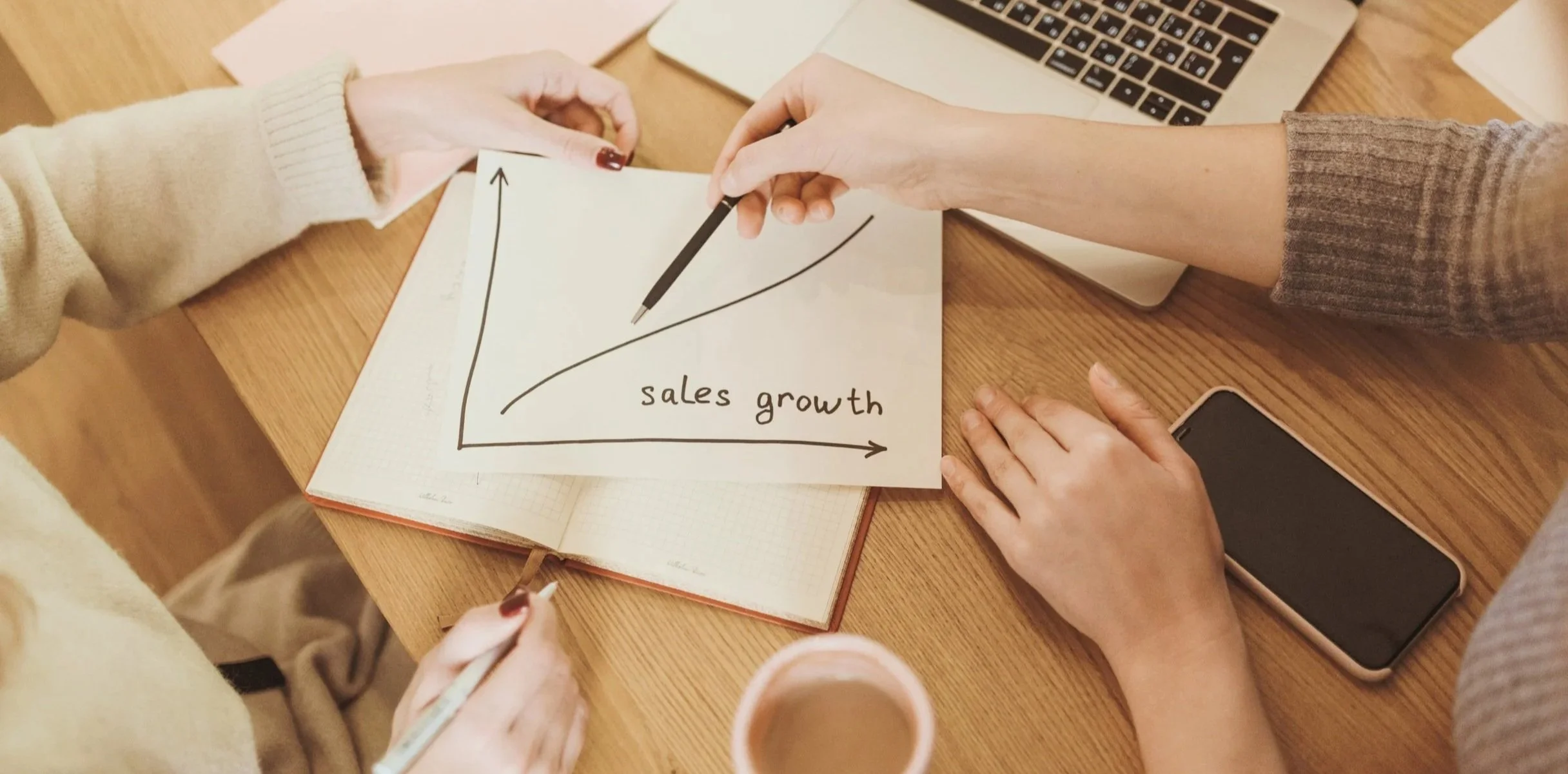 Two people are exchanging a paper with a sales growth graph on it. The workspace includes a laptop, smartphone, notebooks, and a coffee cup.