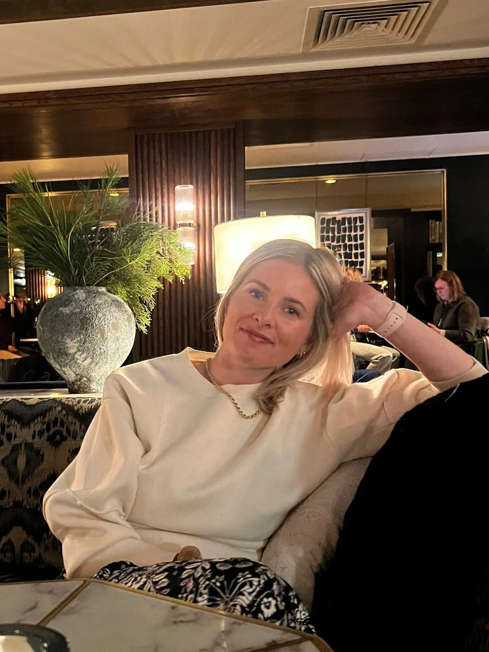 A woman with light-colored hair is sitting at a restaurant, wearing a light-colored top and a gold chain necklace, smiling at the camera. In the background, there are other people, a plant in a stone pot, and warm lighting in a modern, elegant interior.