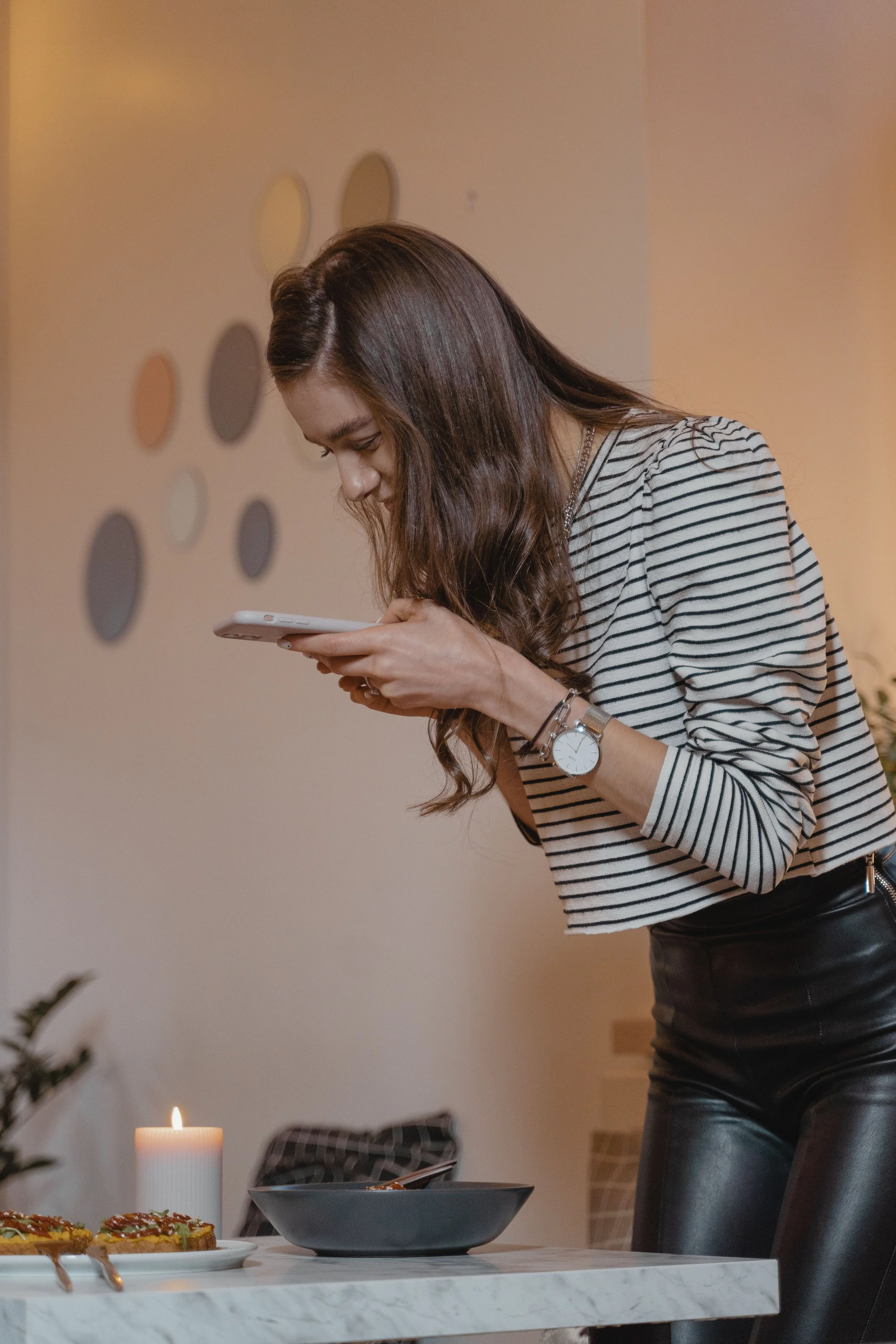 A woman with long brown hair, wearing a striped shirt and black leather pants, looking down at a smartphone in her hands. There is a table with food and a lit candle in the foreground, and a wall with circular decorations in the background.