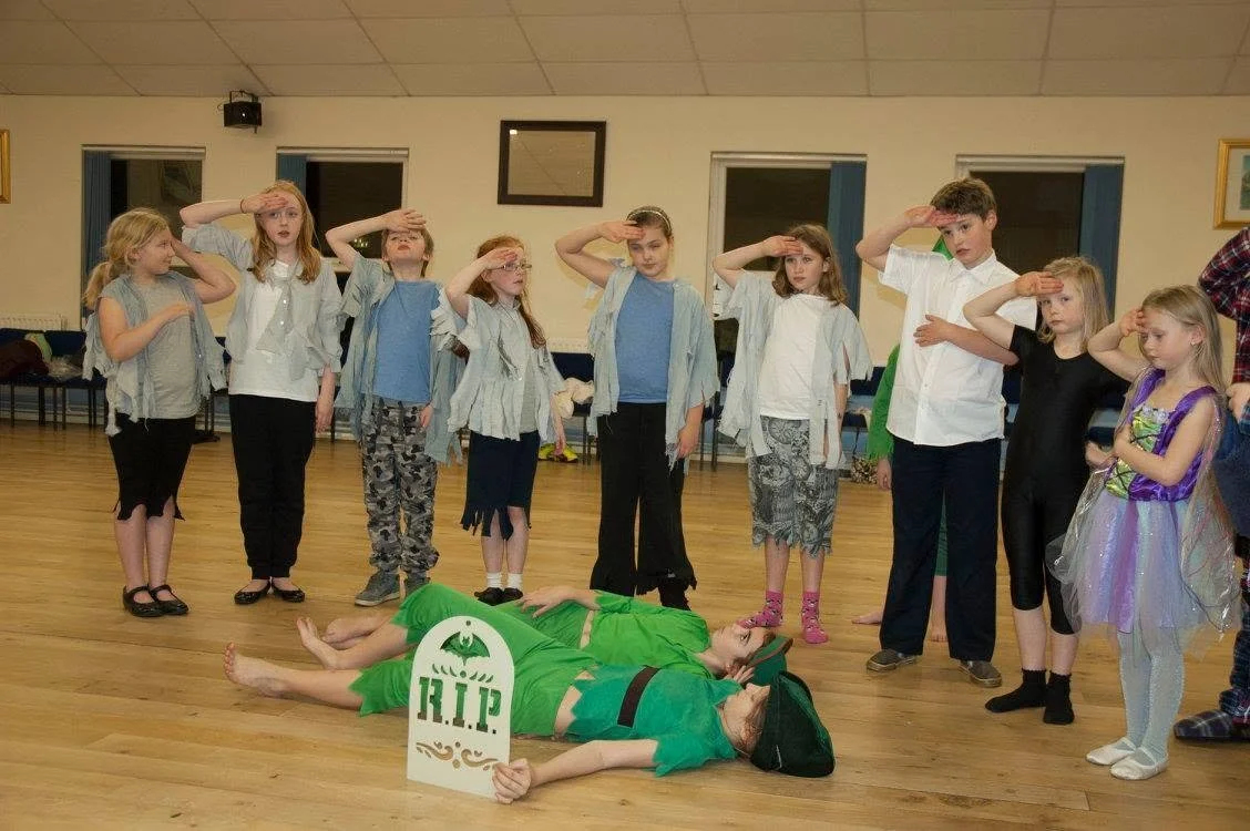 Children dressed as soldiers and characters standing in a line saluting, with two children lying on the floor dressed as a corpse, holding a 'R.I.P' sign, in a room with wooden floors and plain walls.