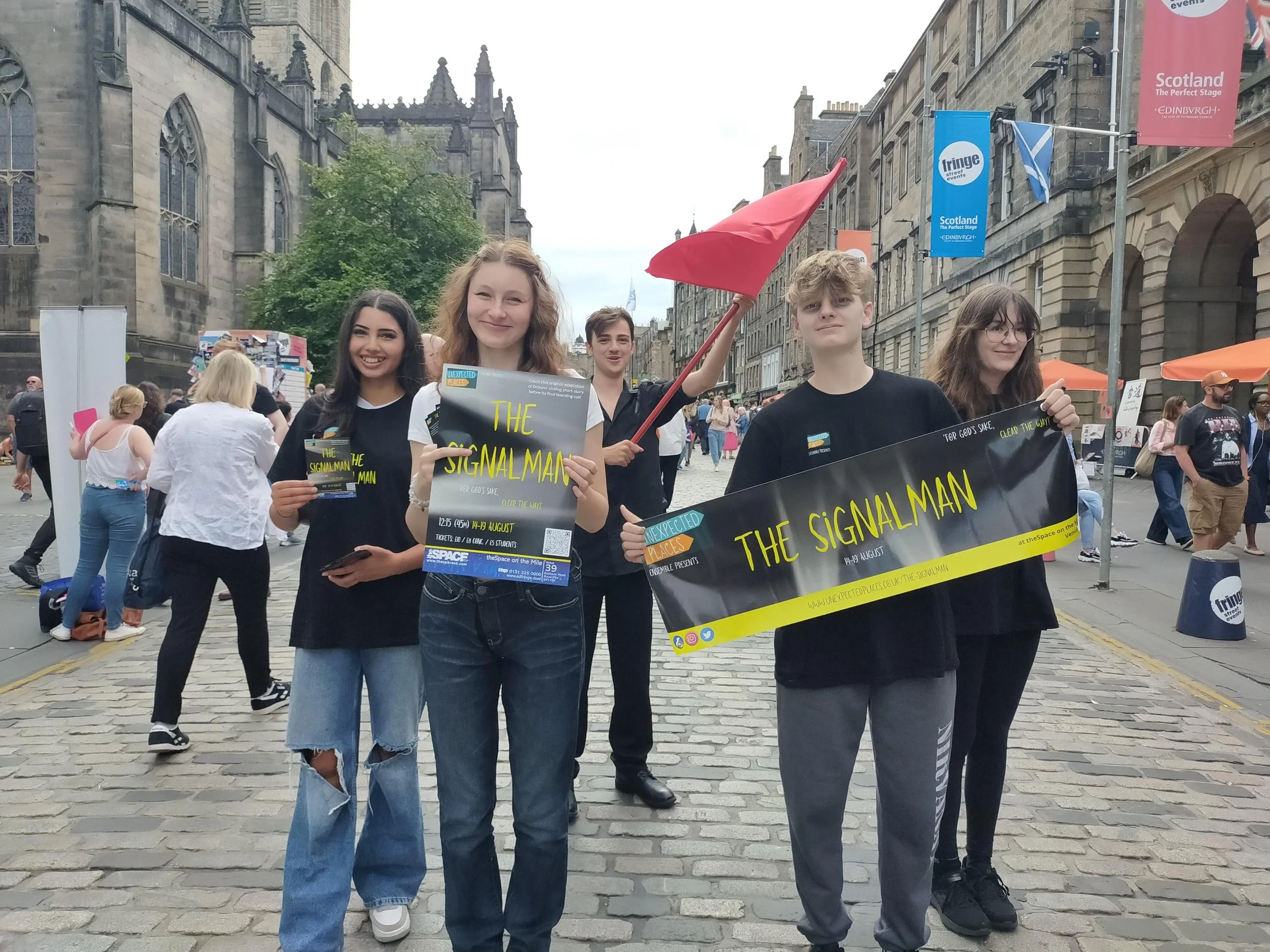 A group of young people participating in a street event, holding signs and banners for a performance called 'The Signal Man' at the Fringe street event in Edinburgh, with historic buildings and other attendees in the background.