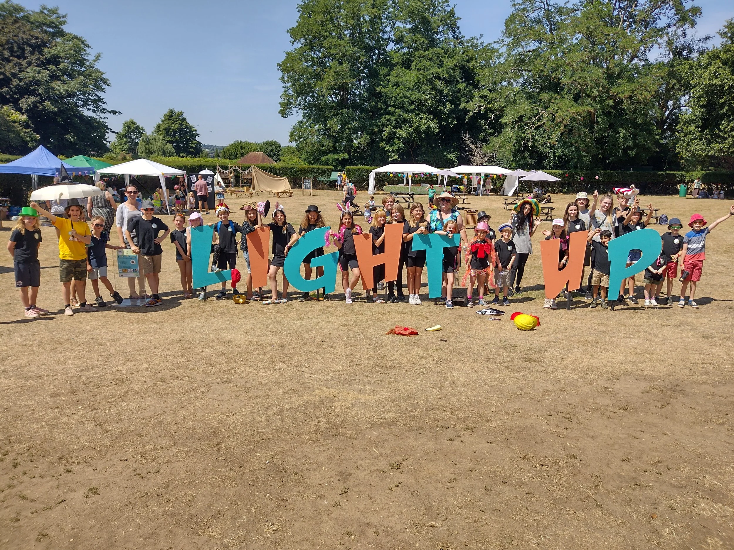 Group of children and adults gathering in a park for an outdoor event with white tents, balloons, and trees in the background, some children holding large colorful letters.