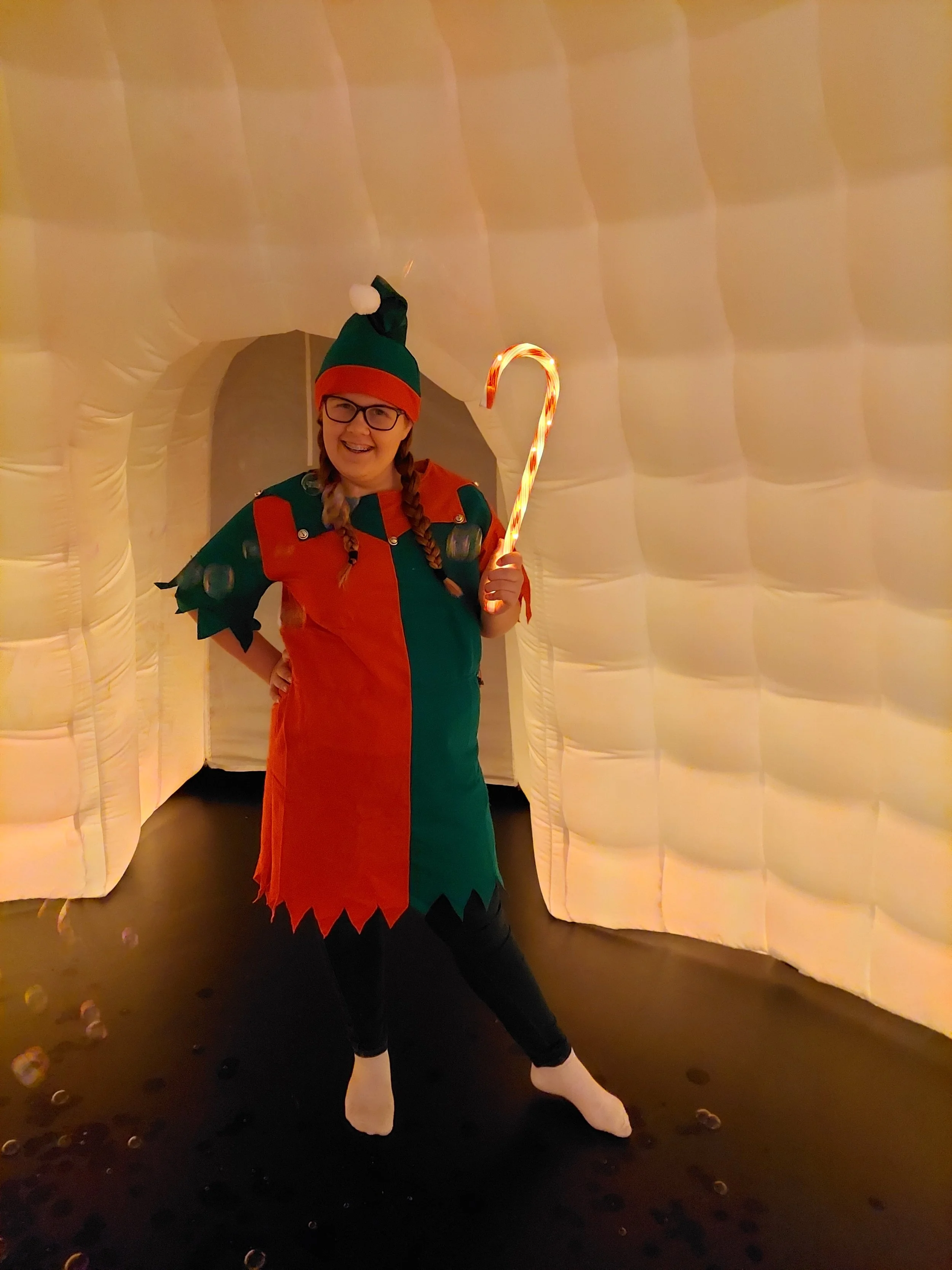 Woman dressed as an elf holding a candy cane, posing in front of an inflatable holiday backdrop.