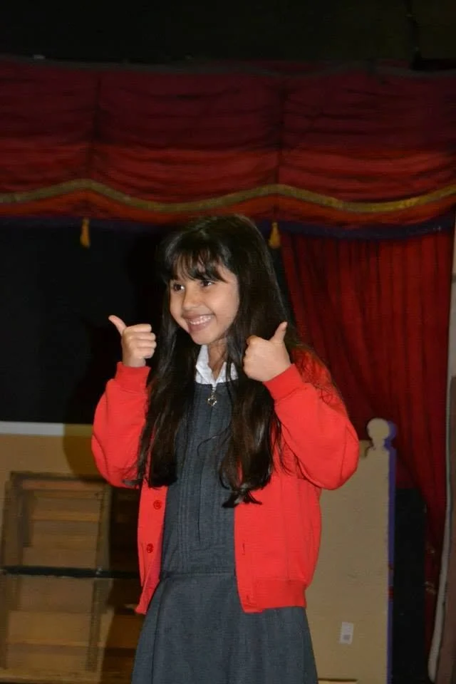 A young girl with long dark hair, wearing a gray school uniform and a red cardigan, smiling and giving two thumbs up in front of red stage curtains.