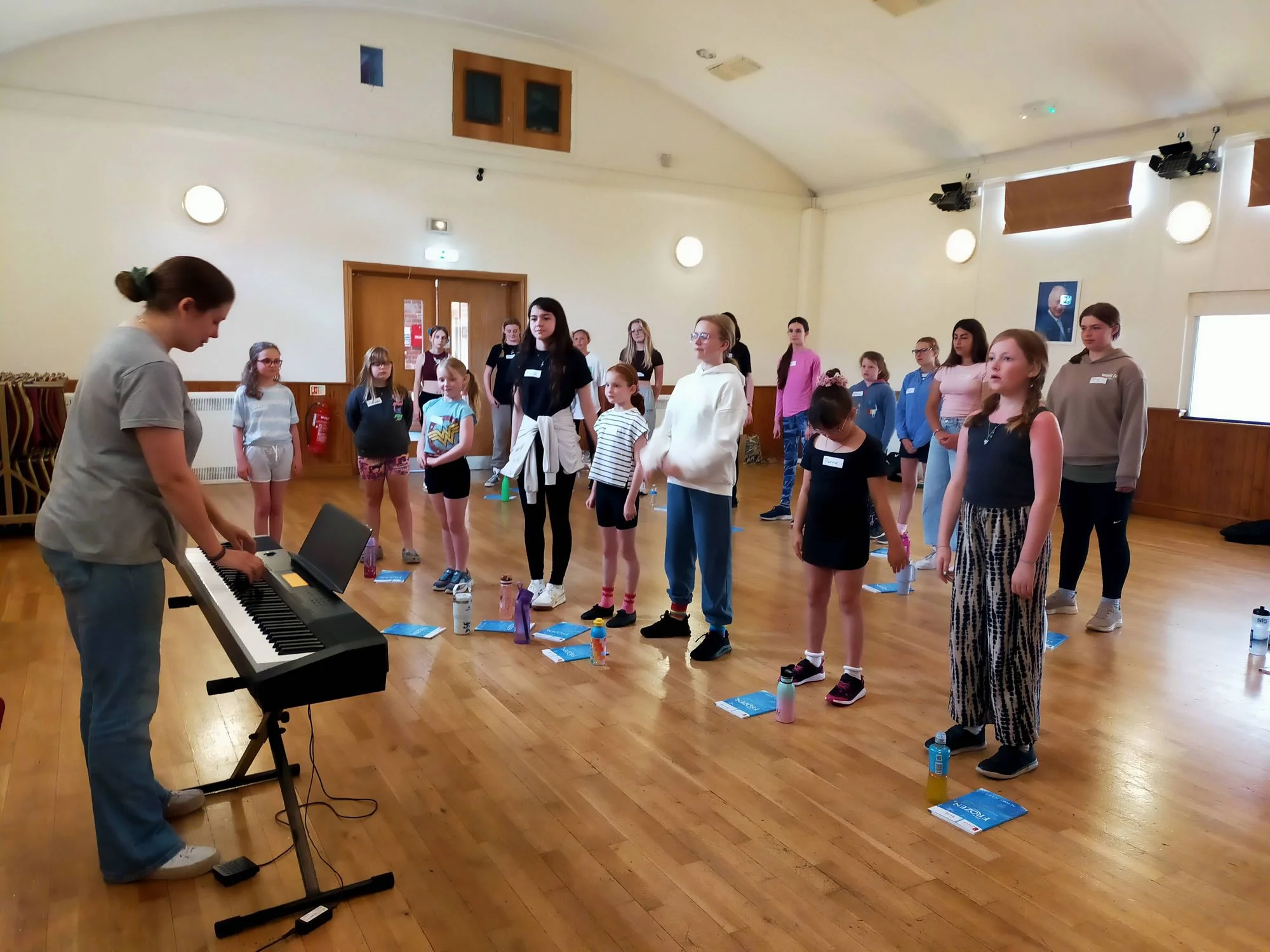 Group of children and teenagers standing in a circle in a spacious room with wooden floors, while a woman plays a keyboard in the foreground.
