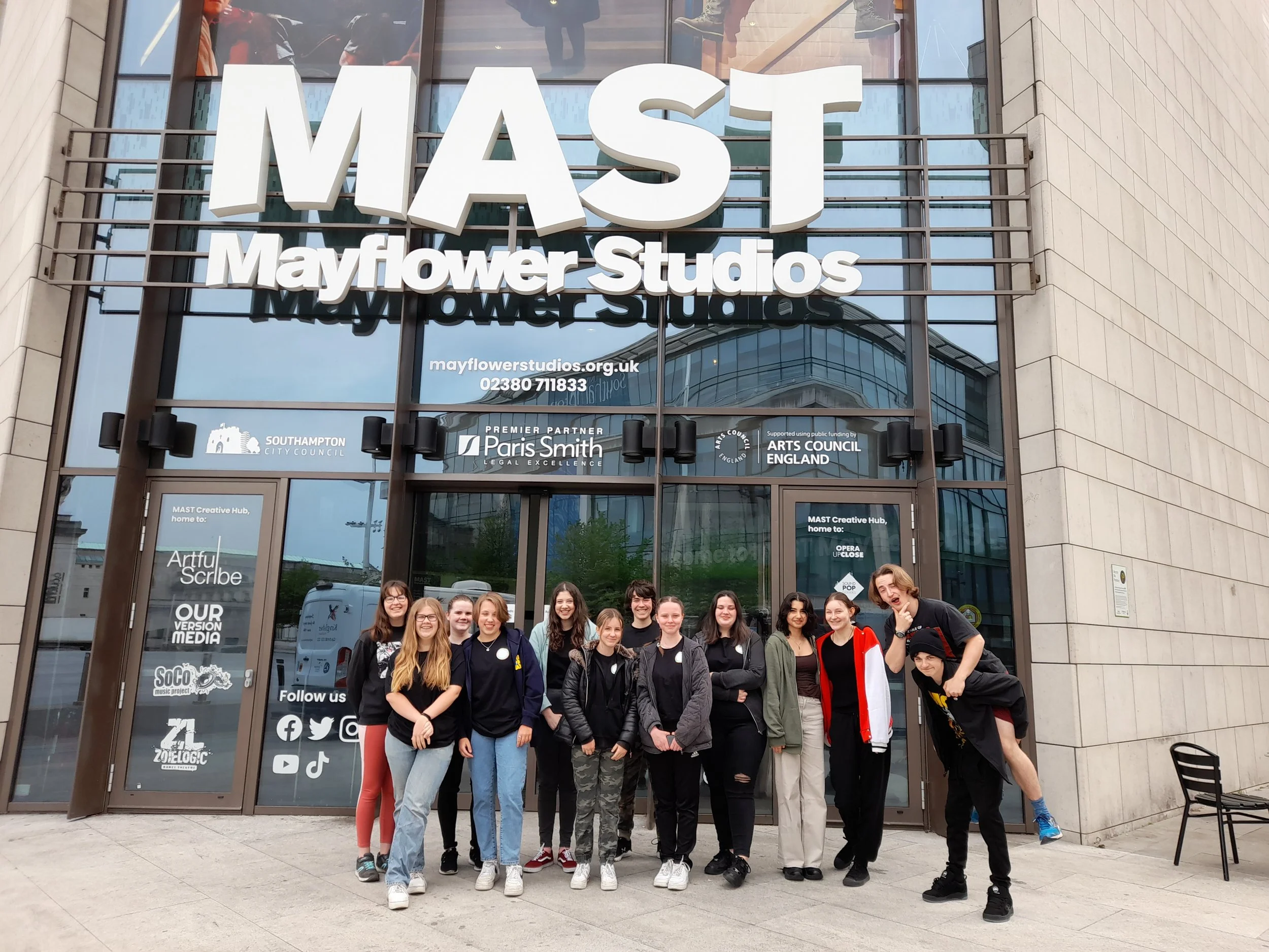 A group of young people standing together in front of the Mayflower Studios building in an urban area, posing for a group photo. The building features a large sign that reads 'MIST Mayflower Studios.' The group appears happy and diverse, with some people smiling, and their reflections can be seen in the glass windows behind them.