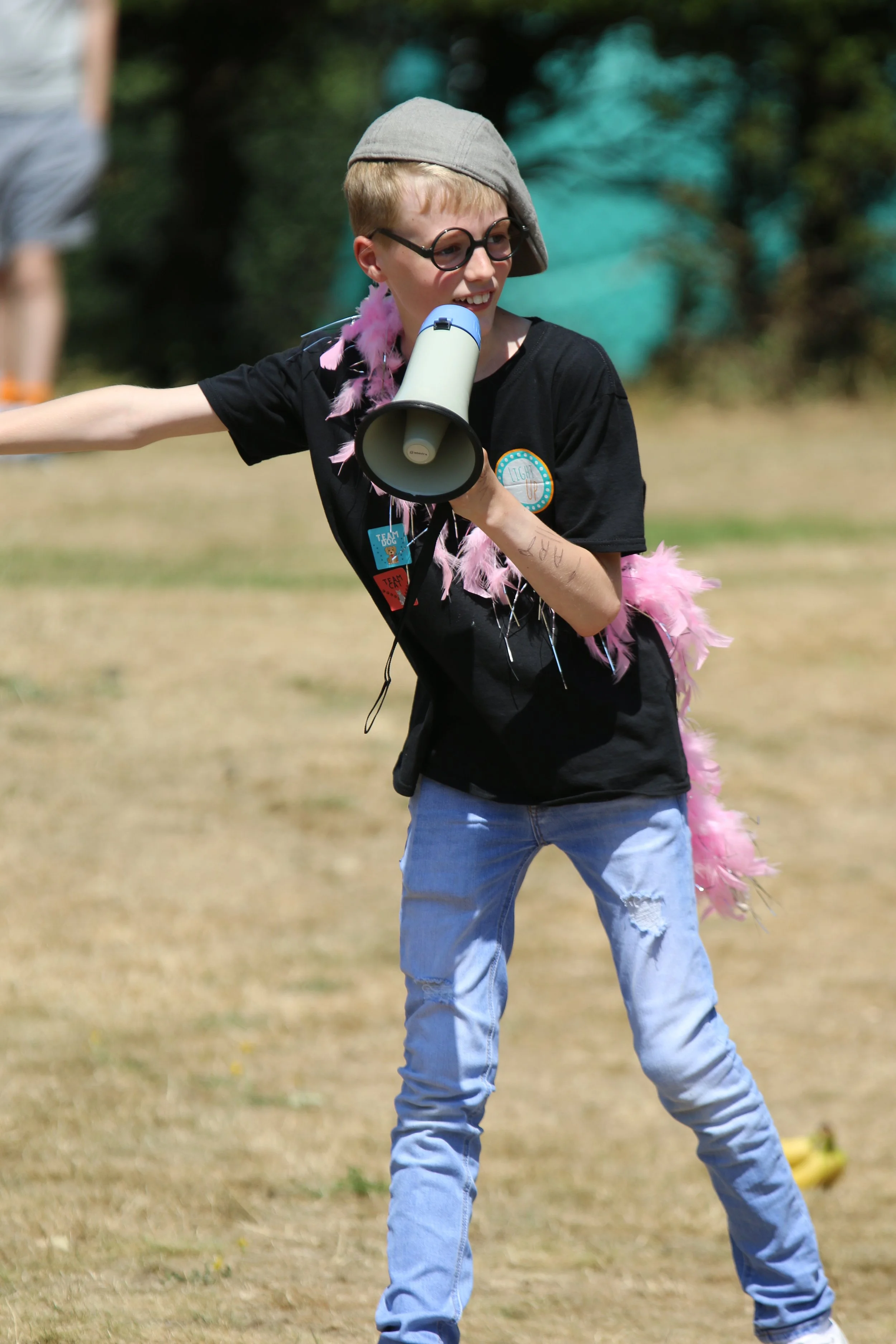 A young person with short blonde hair, wearing glasses, a gray cap, a black T-shirt, and ripped jeans, is outdoors holding a megaphone and wearing pink feather decorations.