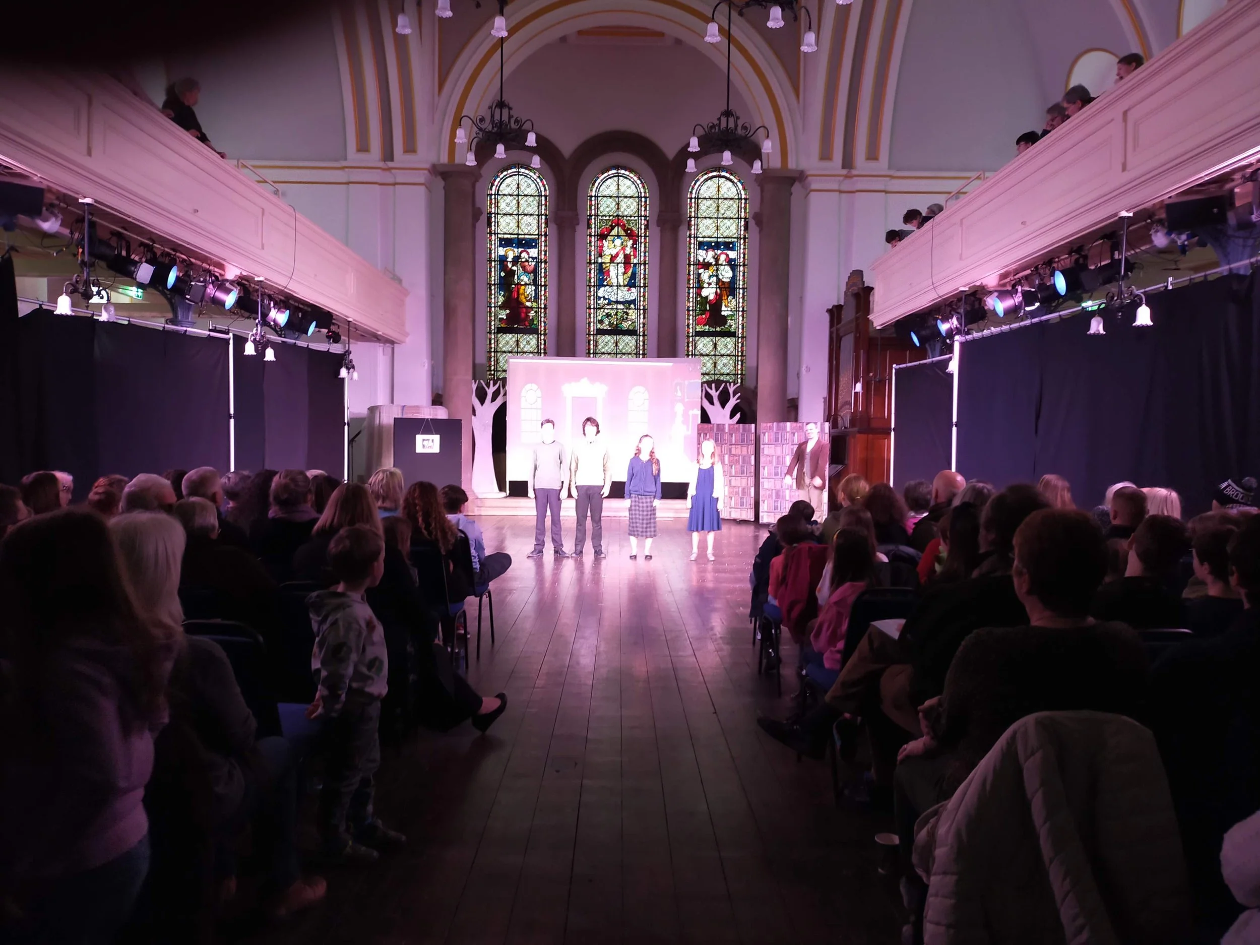 A church hall with stained glass windows at the back, set up for a performance or presentation, with a stage and an audience seated in front. There are people standing on stage.