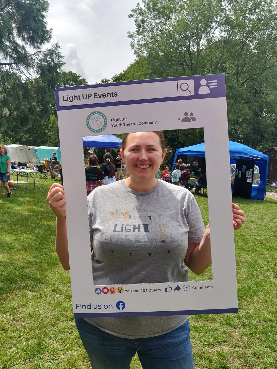 A woman smiling and holding a Facebook-themed photo frame at an outdoor event with tents and people in the background.