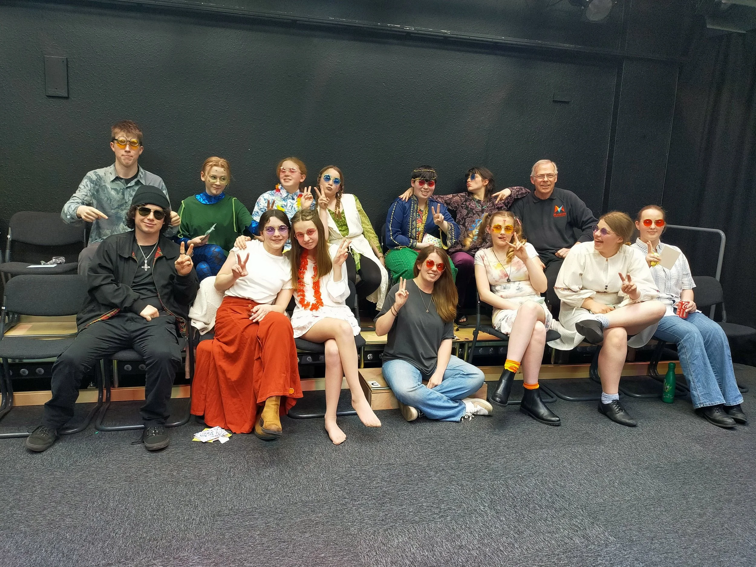 Group of children and an adult in fun costumes and sunglasses, sitting and standing on chairs in a classroom or theater setting, making peace signs and smiling.
